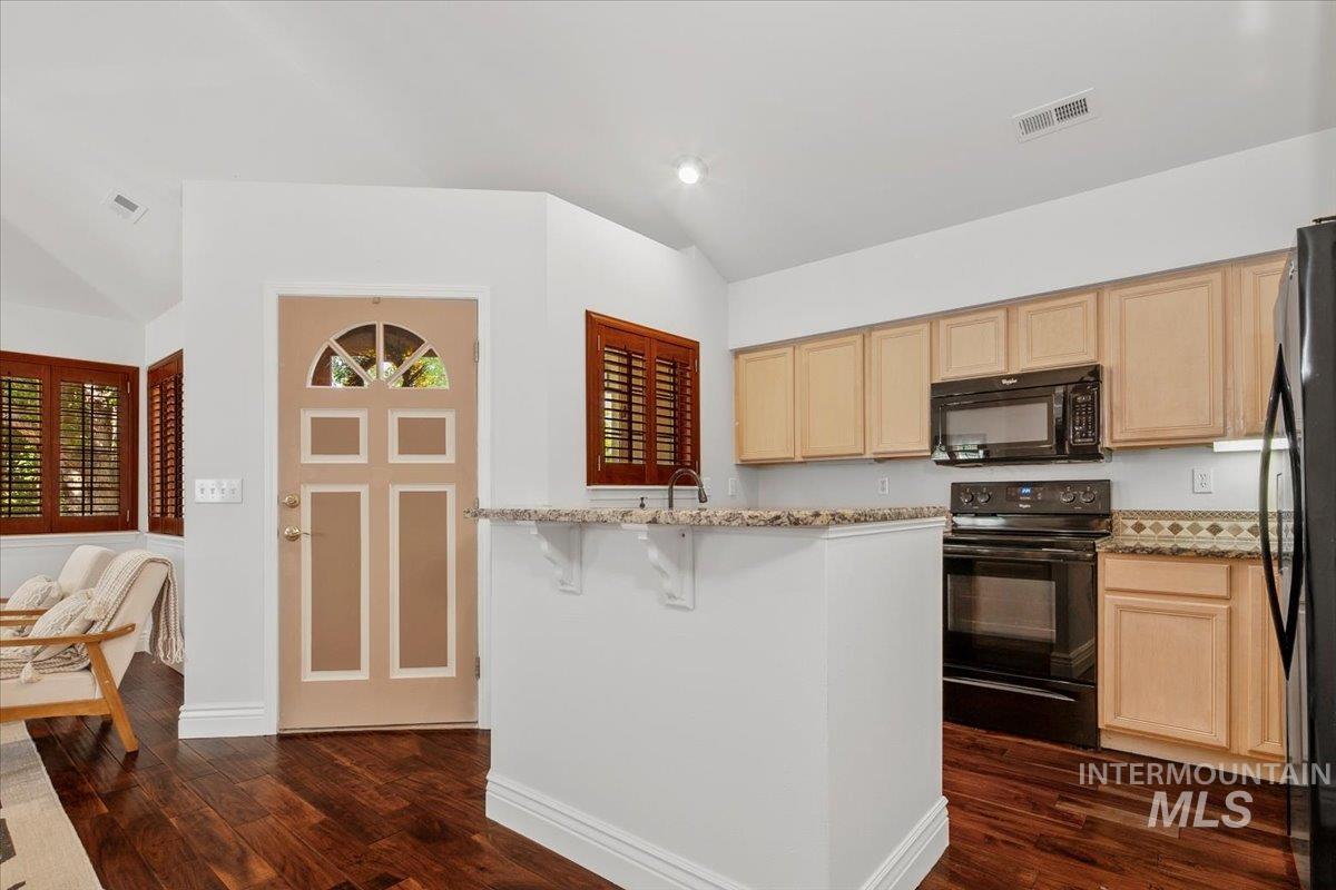 Kitchen featuring black appliances, granite counters, vaulted ceiling, light brown cabinetry, and dark wood finished floors