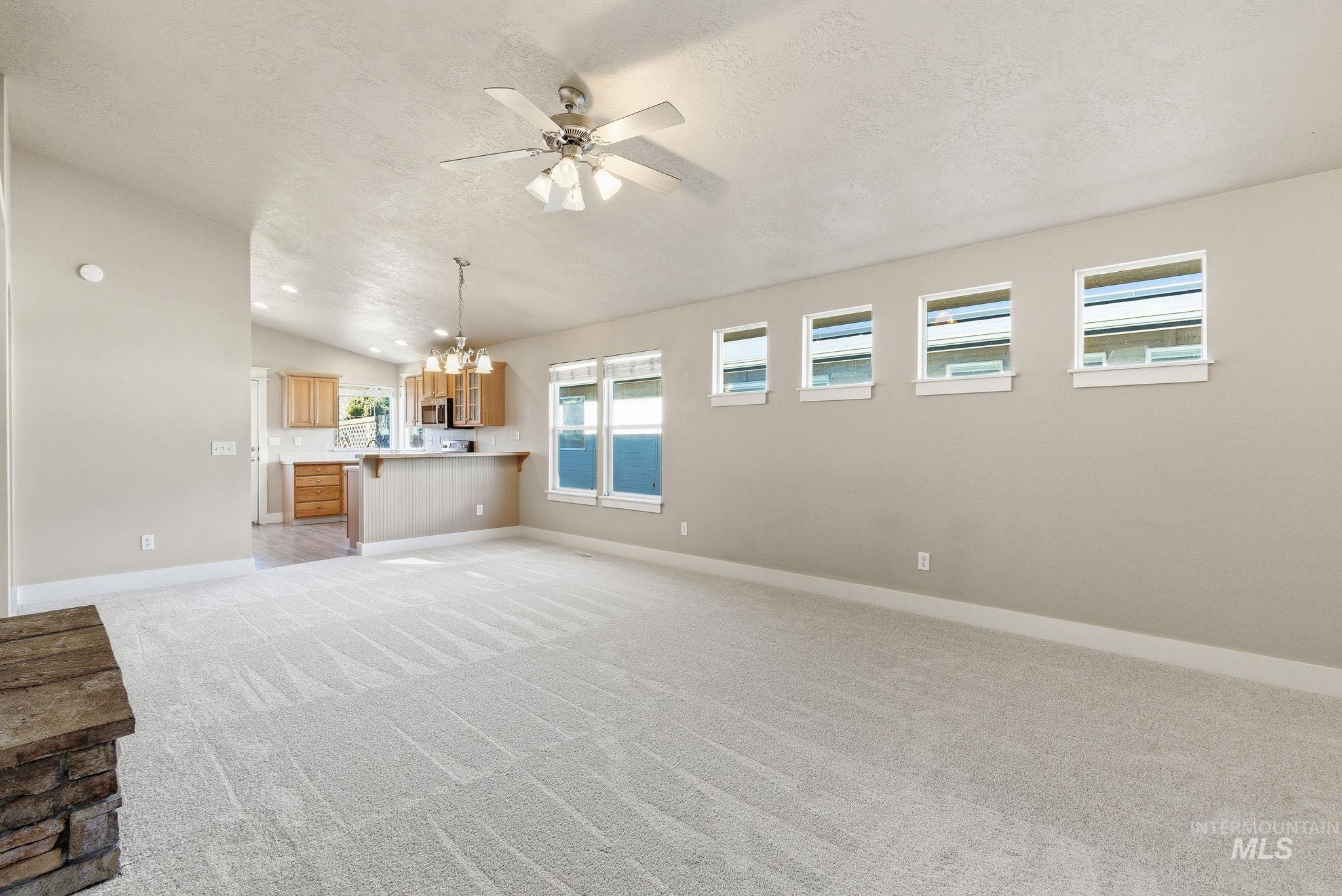 Unfurnished living room with light carpet, a ceiling fan, vaulted ceiling, a chandelier, and a textured ceiling
