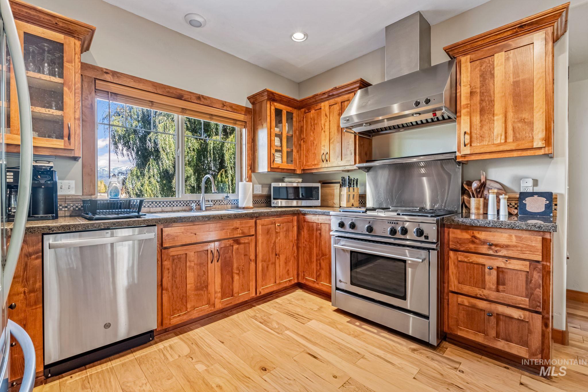 Kitchen with wall chimney range hood, stainless steel appliances, glass insert cabinets, light wood-type flooring, and brown cabinets