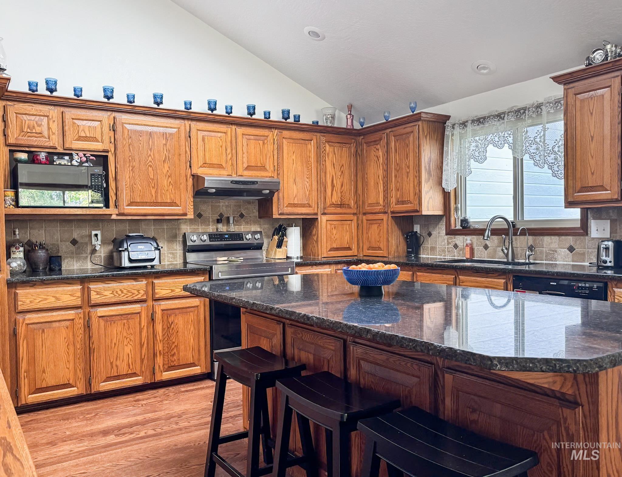 Kitchen with decorative backsplash, lofted ceiling, brown cabinets, light wood-style floors, and dark stone counters