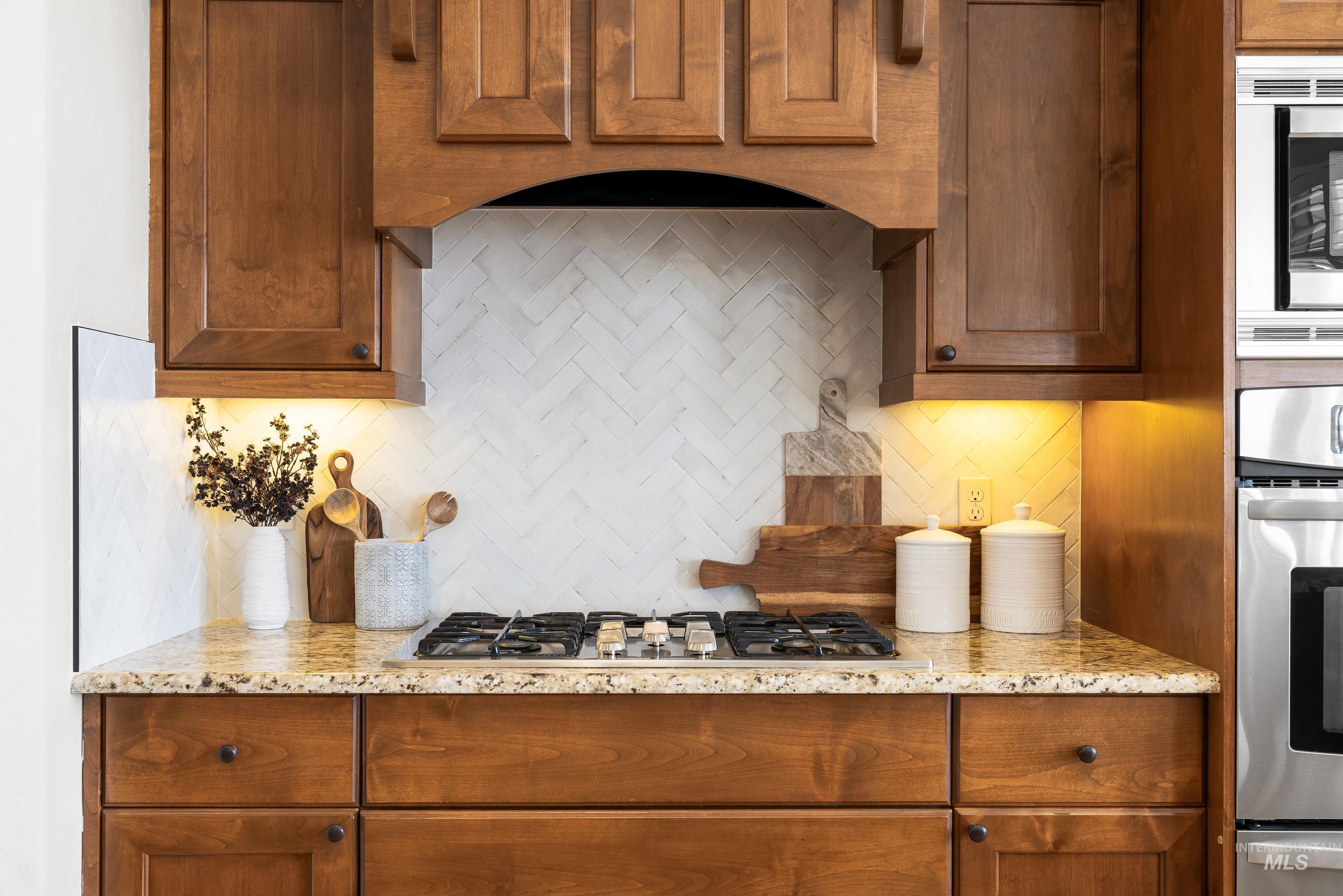 Kitchen featuring backsplash, brown cabinets, light stone counters, ventilation hood, and stainless steel appliances