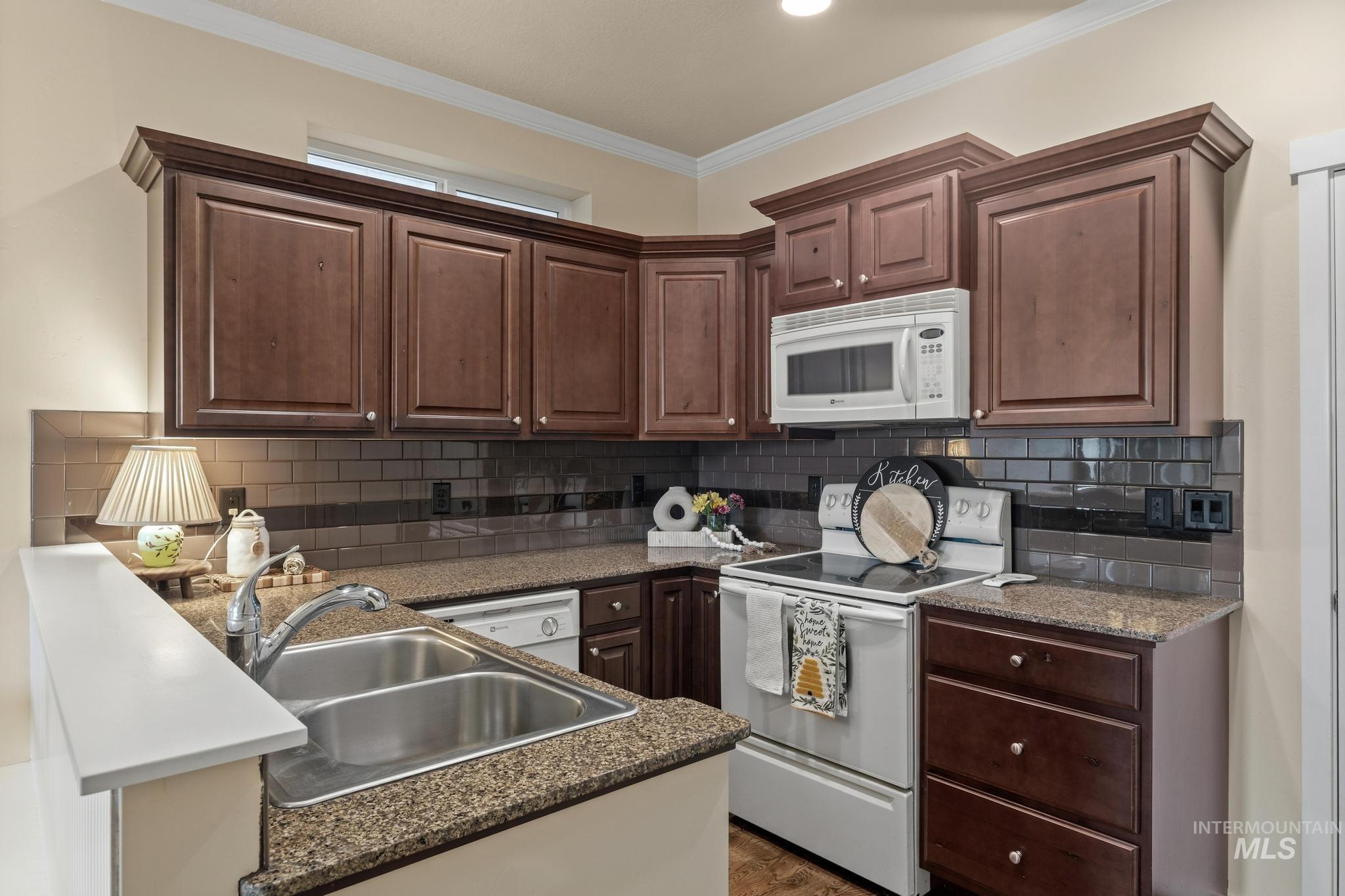 Kitchen with white appliances, dark wood finish cabinetry, ornamental molding, a peninsula, and tasteful backsplash