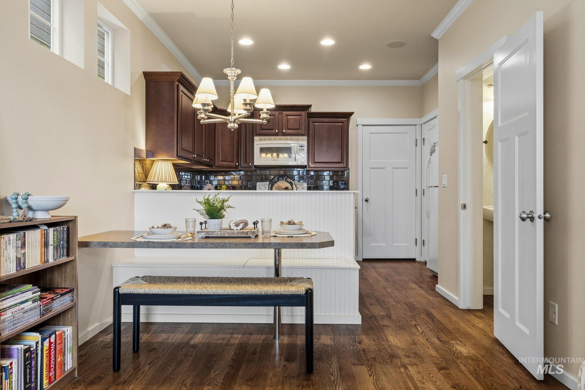 Kitchen featuring dark wood finish cabinets, backsplash, white appliances, suspended lighting, and crown molding