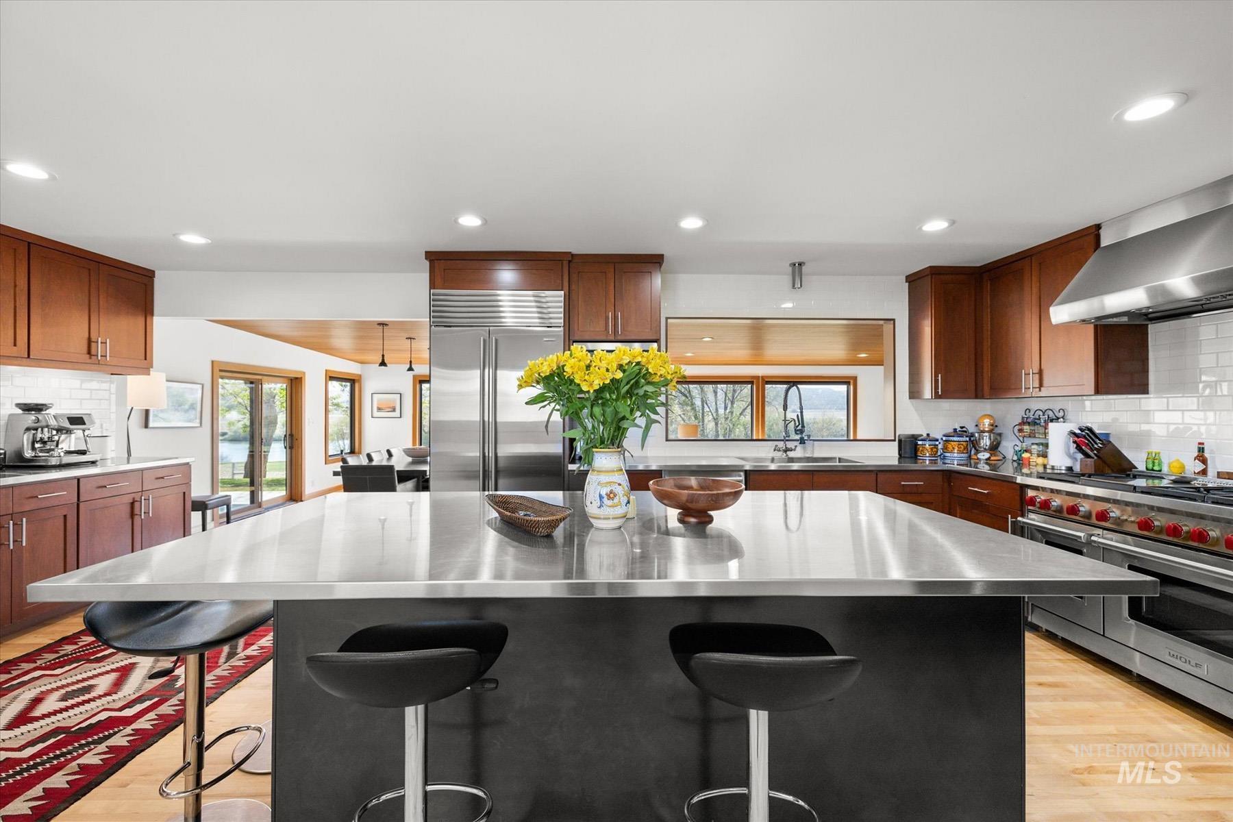Kitchen featuring stainless steel countertops, high quality appliances, wall chimney exhaust hood, backsplash, and light wood finished floors