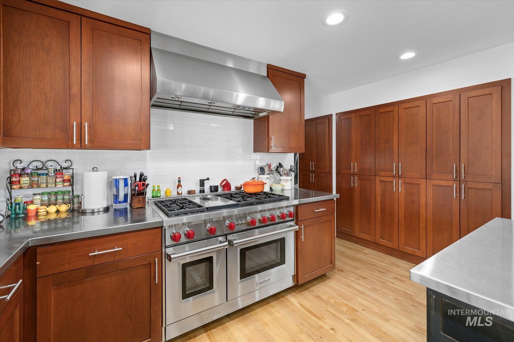 Kitchen with stainless steel counters, wall chimney exhaust hood, range with two ovens, decorative backsplash, and light wood-style floors