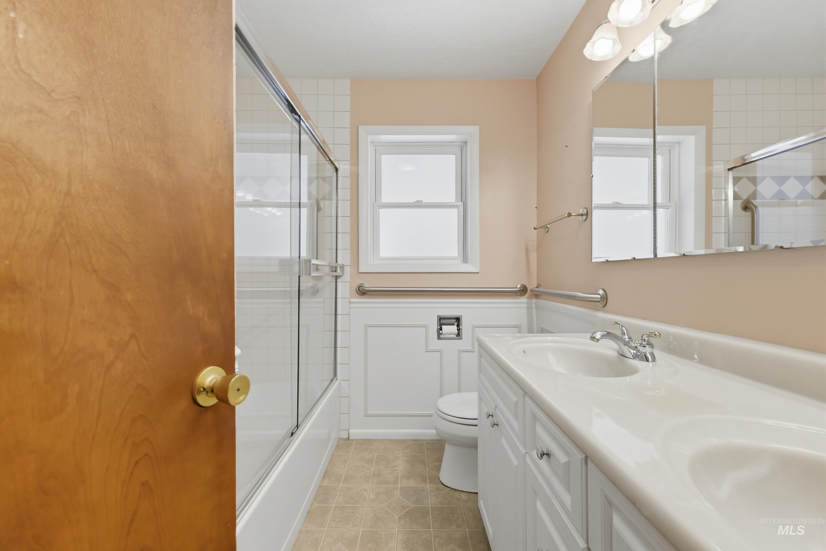 Bathroom featuring a double vanity with integrated sinks, a tub/shower combination with glass enclosure, white wainscoting, and a window