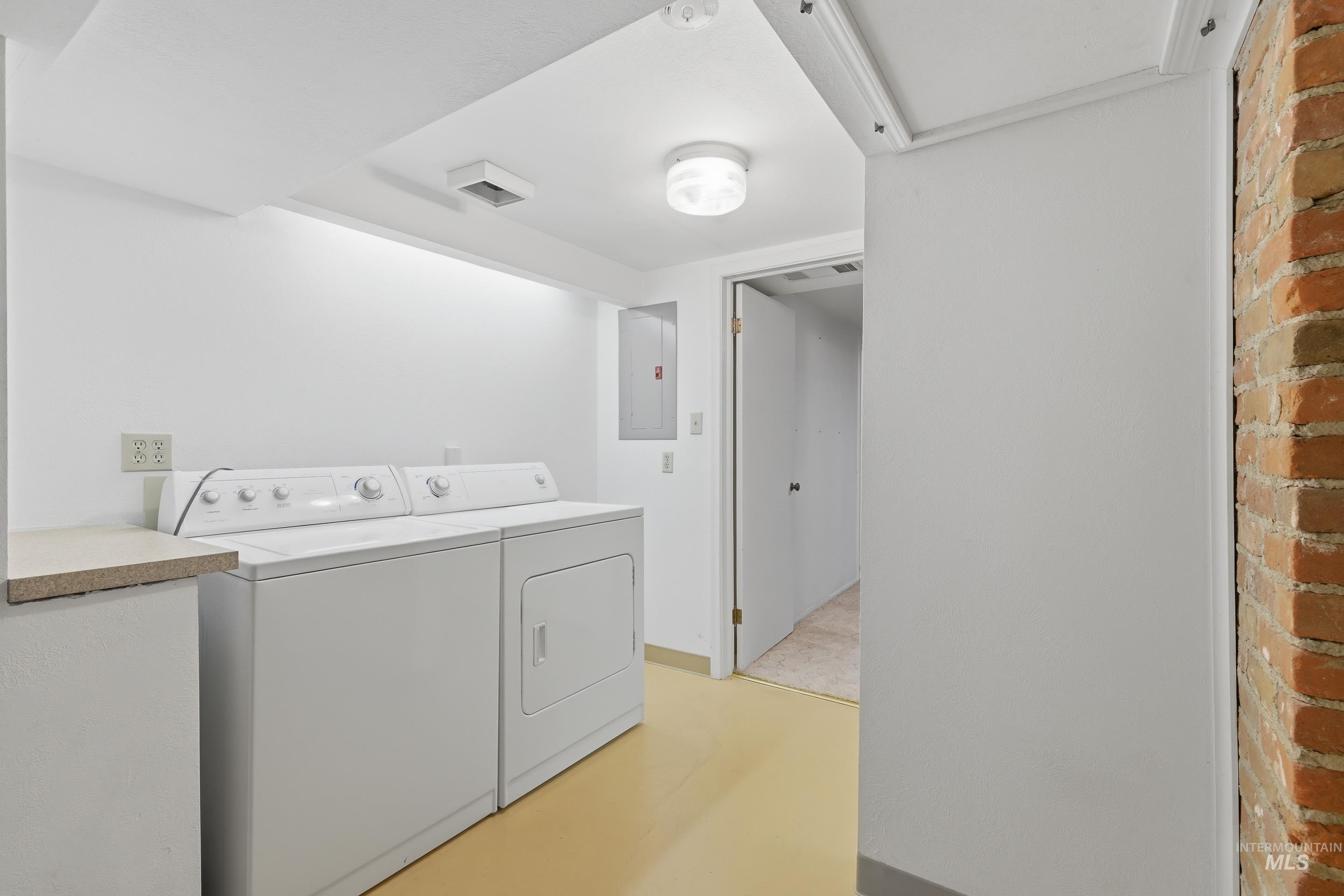 Dedicated laundry area featuring a washer and dryer, light-toned flooring, exposed brick wall, and overhead lighting