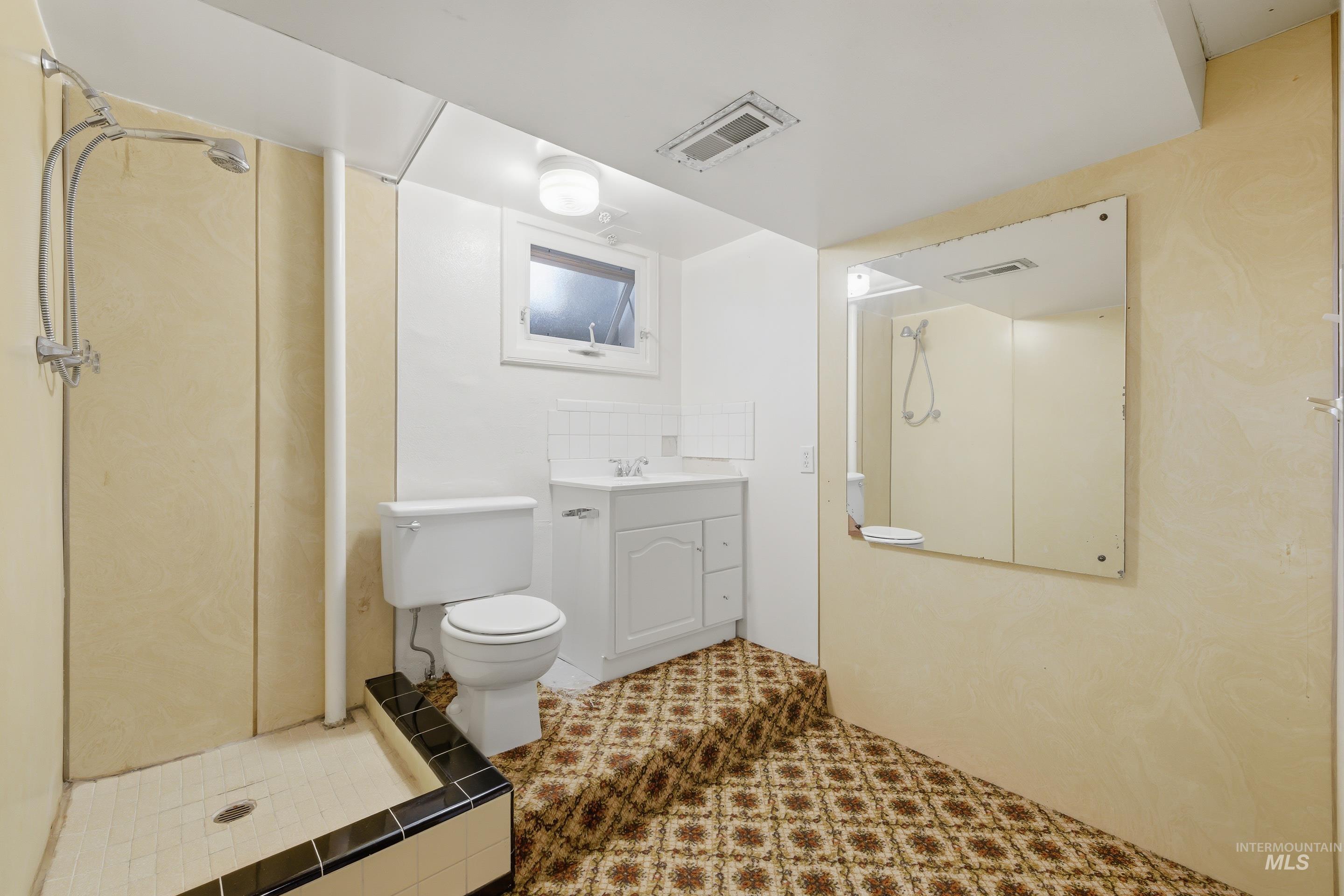 Bathroom featuring a standing shower with a tiled base and dark trim, a white vanity with a single sink, a toilet, a window, and a wall-mounted mirror