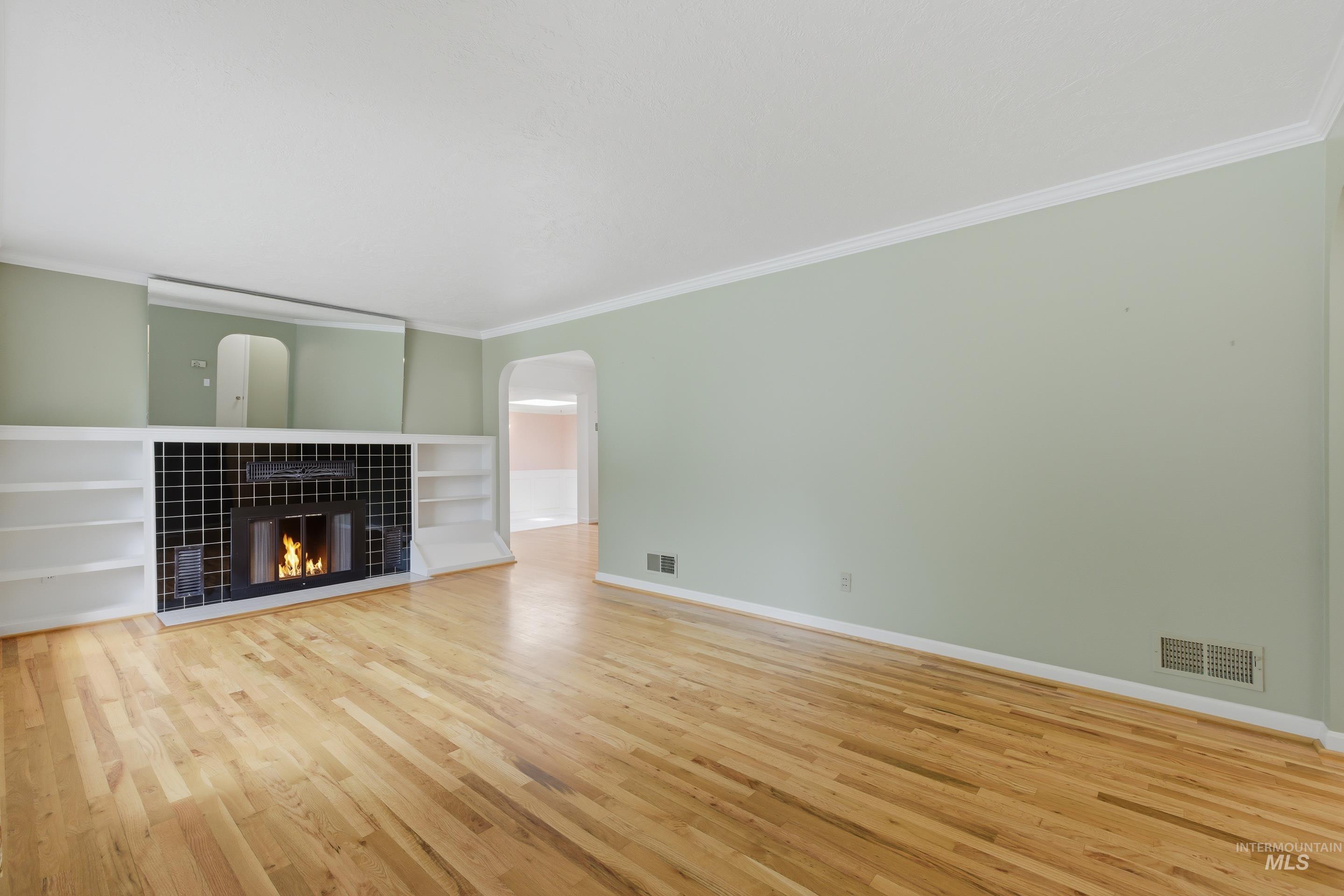 Living area featuring light wood-finish flooring, a fireplace with black tile surround, built-in shelving, white crown molding, and an arched doorway