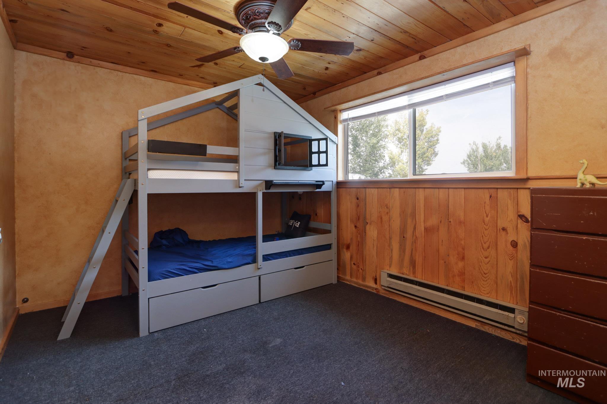 Unfurnished bedroom with a baseboard radiator, a ceiling fan, wood ceiling, wood walls, and dark colored carpet