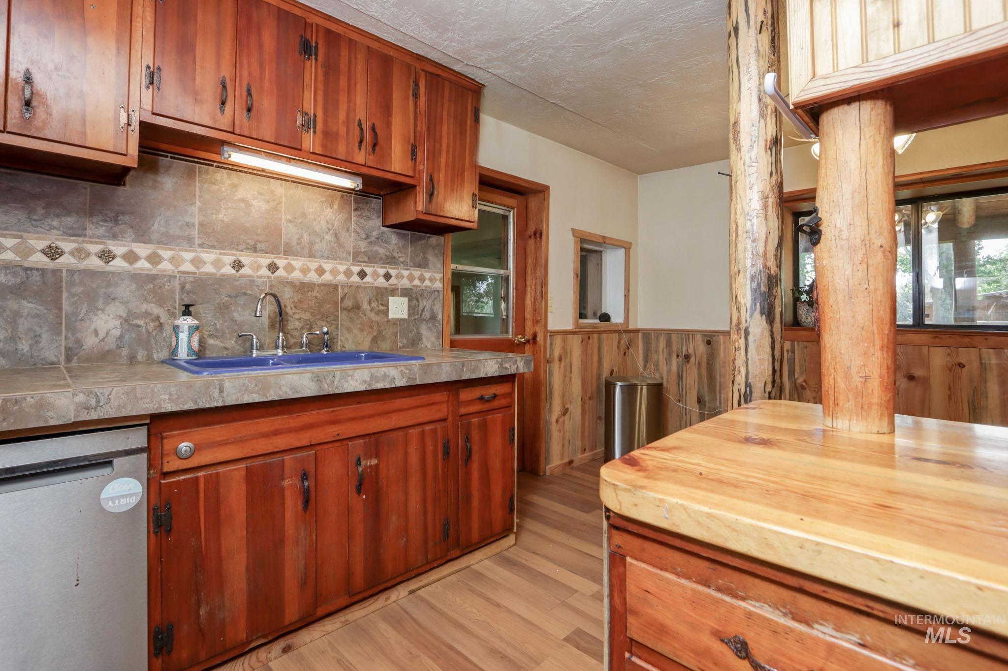 Kitchen featuring light countertops, stainless steel dishwasher, light wood-style flooring, brown cabinets, and wainscoting