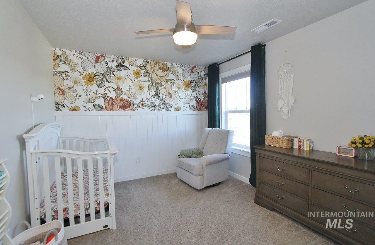 Bedroom with wainscoting, wallpapered walls, a nursery area, ceiling fan, and light carpet