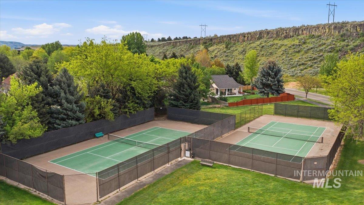 Two fenced tennis courts with green playing surfaces, surrounded by mature trees and natural landscaping