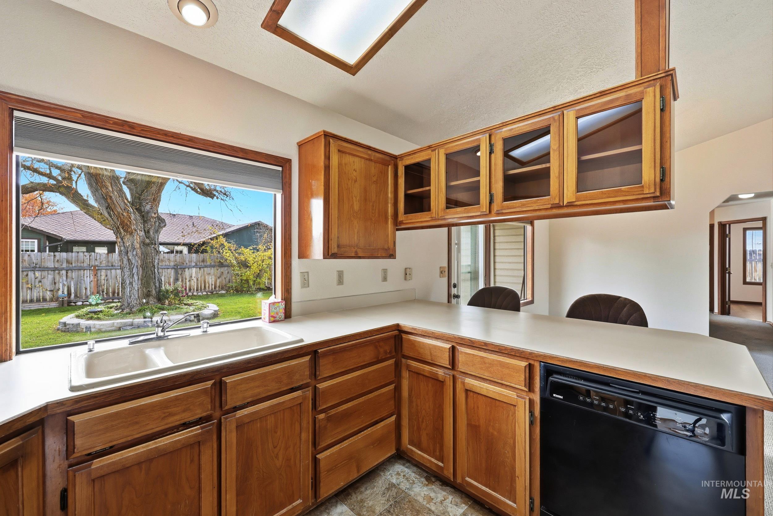 Kitchen featuring brown cabinets, black dishwasher, light countertops, glass insert cabinets, and vaulted ceiling