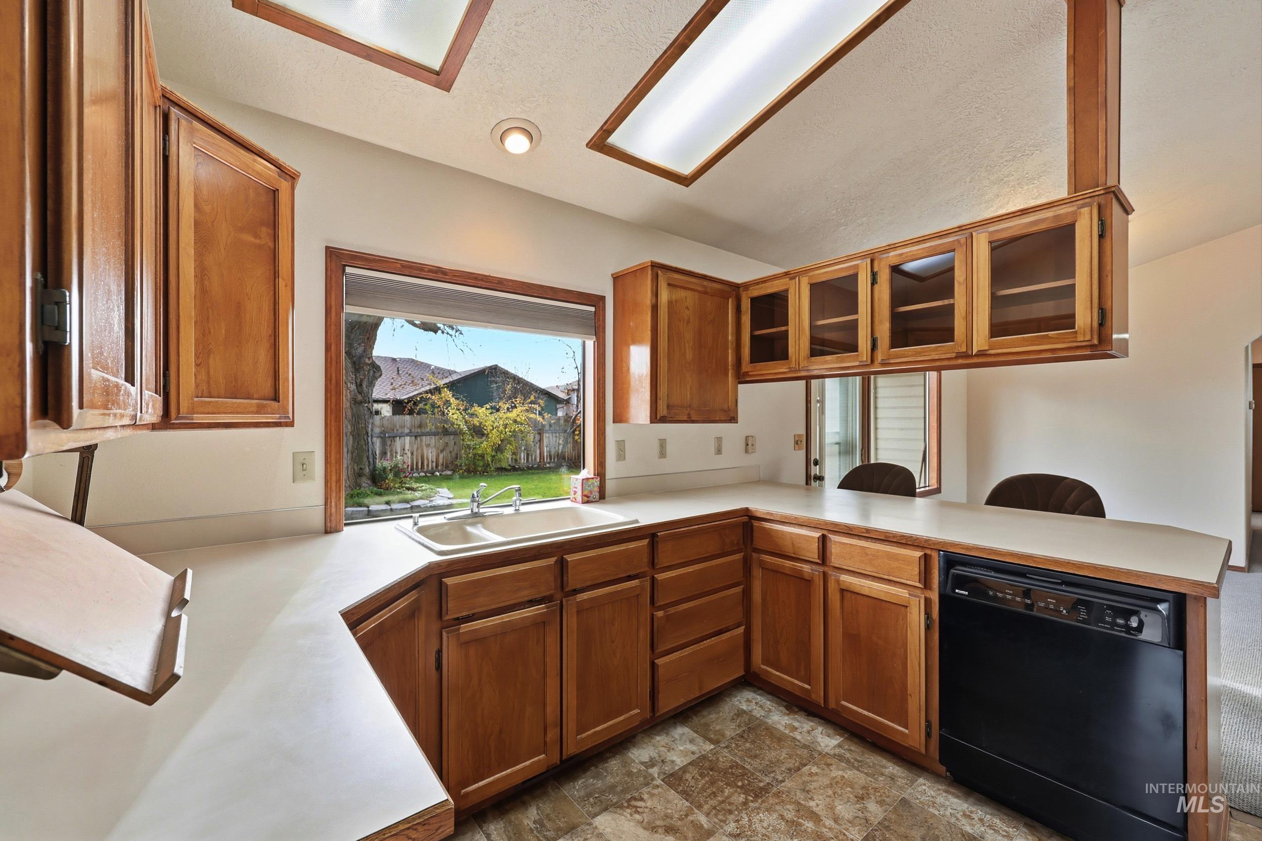 Kitchen with brown cabinetry, light countertops, dishwasher, a textured ceiling, and glass insert cabinets