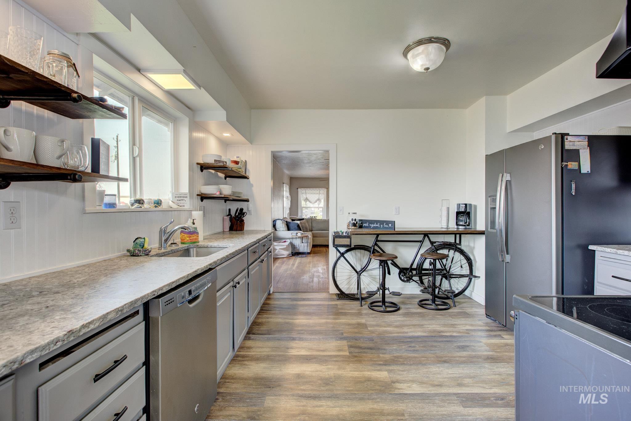 Kitchen featuring wood-finish flooring, light-colored countertops, a stainless steel sink, a stainless steel dishwasher, and open shelving