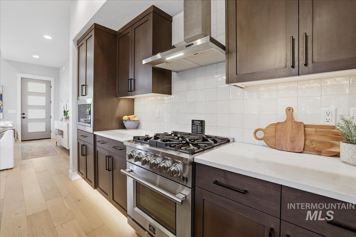 Kitchen featuring dark brown cabinets, stainless steel appliances, wall chimney range hood, tasteful backsplash, and light wood finished floors
