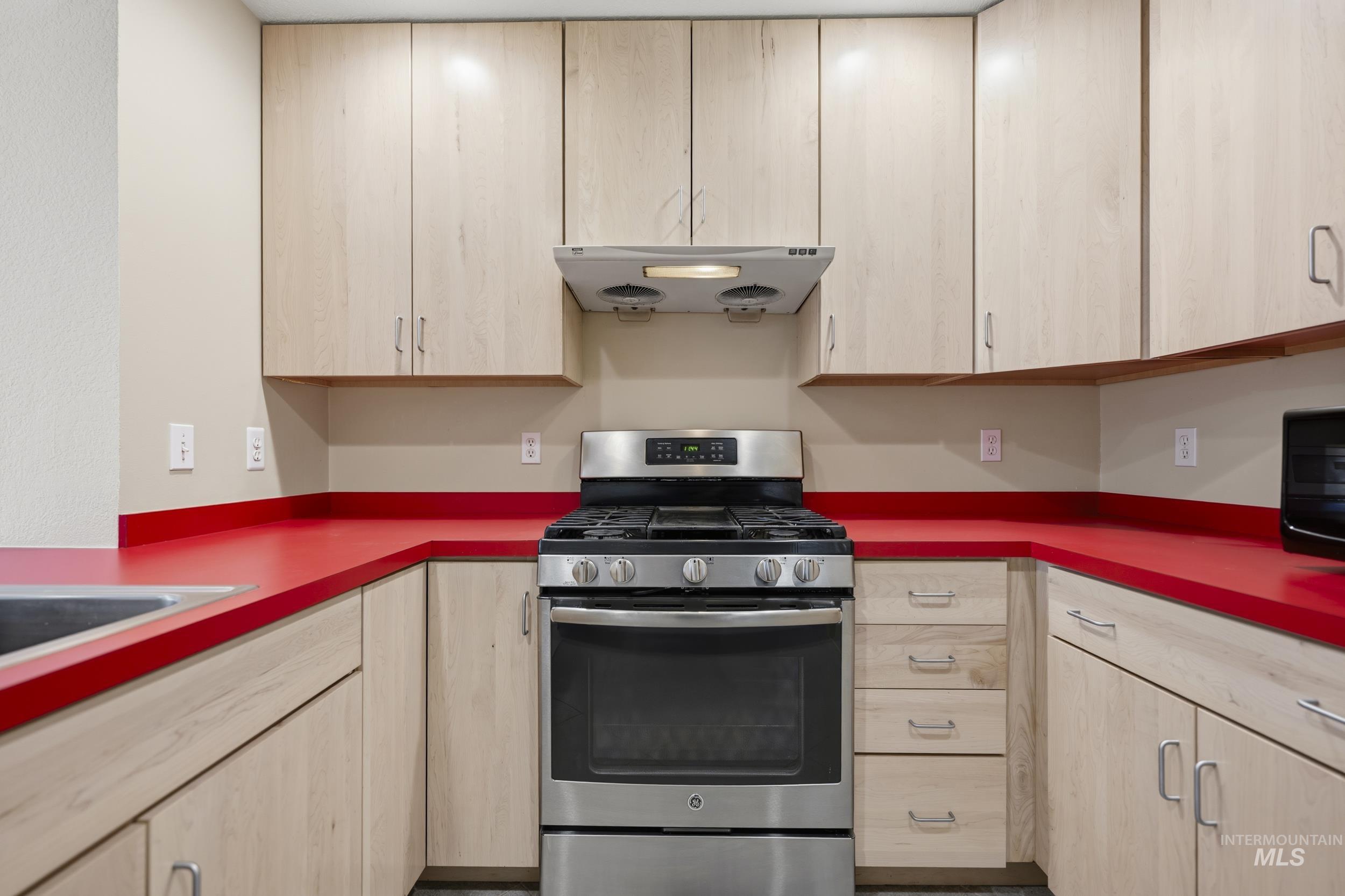 Kitchen with gas range, light wood finish cabinets, ventilation hood, and dark countertops