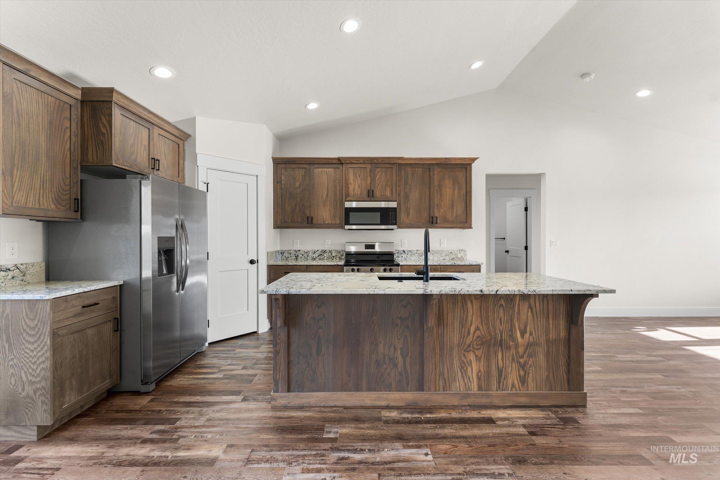 Kitchen with light stone countertops, stainless steel appliances, lofted ceiling, a center island with sink, and dark wood-style flooring