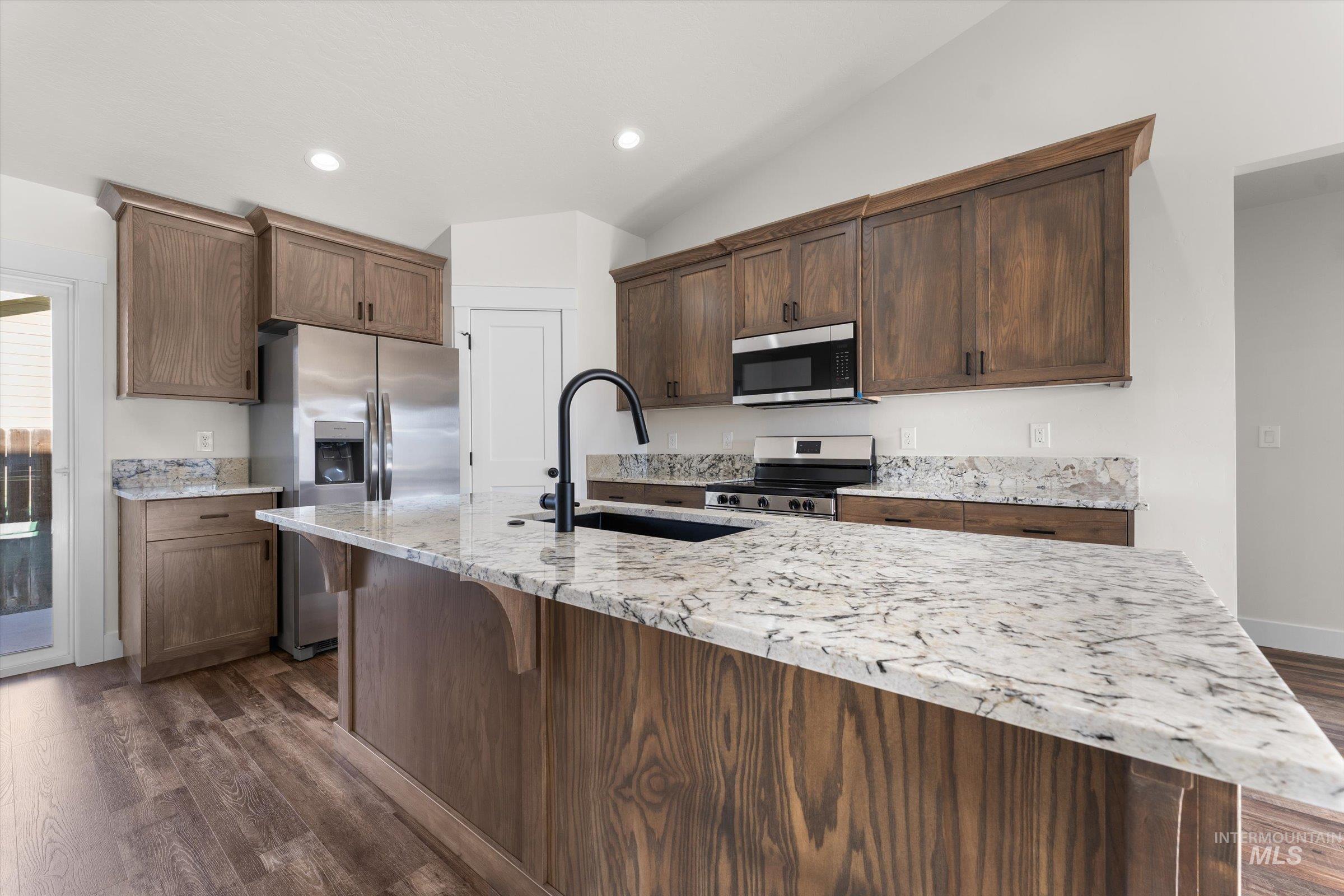 Kitchen featuring light stone countertops, stainless steel appliances, an island with sink, dark wood-style flooring, and lofted ceiling