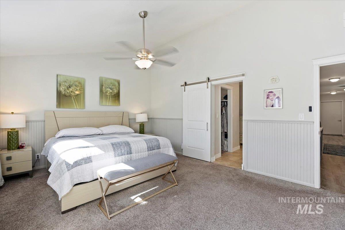 Bedroom featuring vaulted ceiling, a barn door, carpet, wainscoting, and a ceiling fan