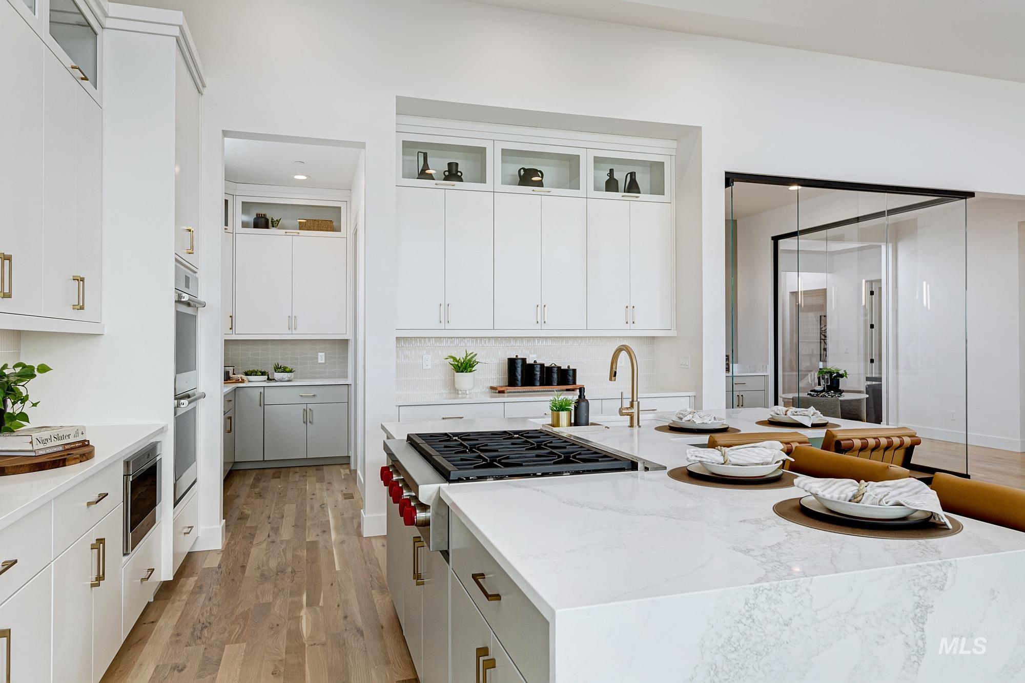 Spacious kitchen featuring a large island with a built-in cooktop, light-toned wood-finish flooring, white cabinetry with brass hardware, and integrated double ovens