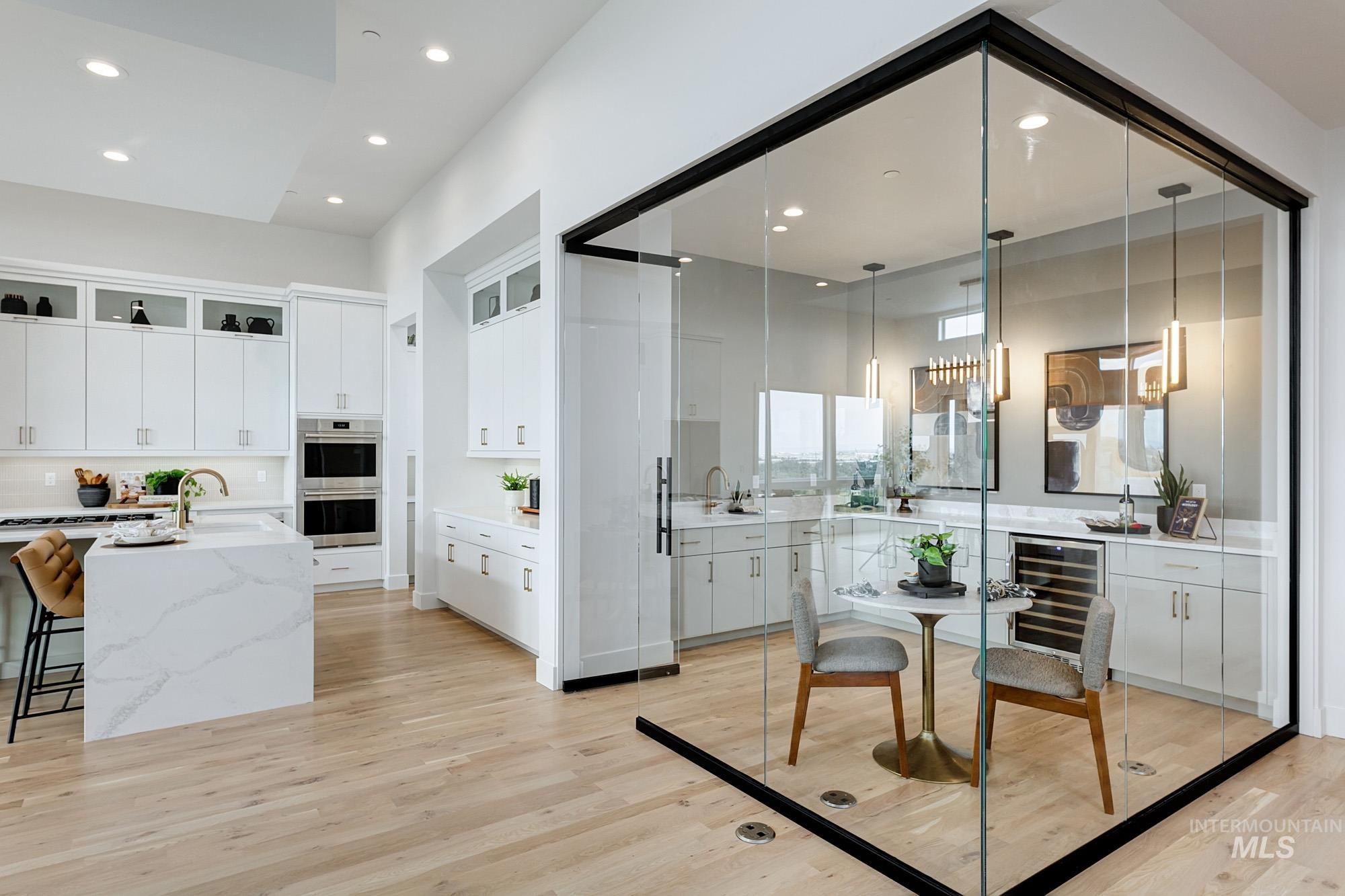 Open-concept kitchen featuring an oversized island with waterfall countertop, integrated double ovens, and a dedicated beverage bar with a wine refrigerator, all complemented by light wood-finish flooring
