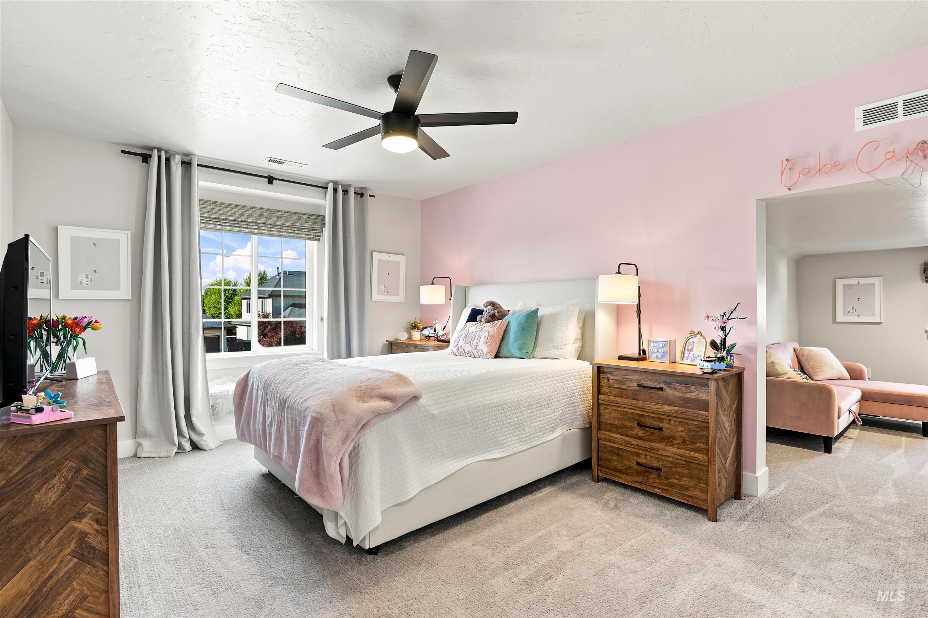 Spacious bedroom featuring light neutral carpet, a large window with roller shades and curtains, a ceiling fan with integrated lighting, and a recessed opening to an additional room