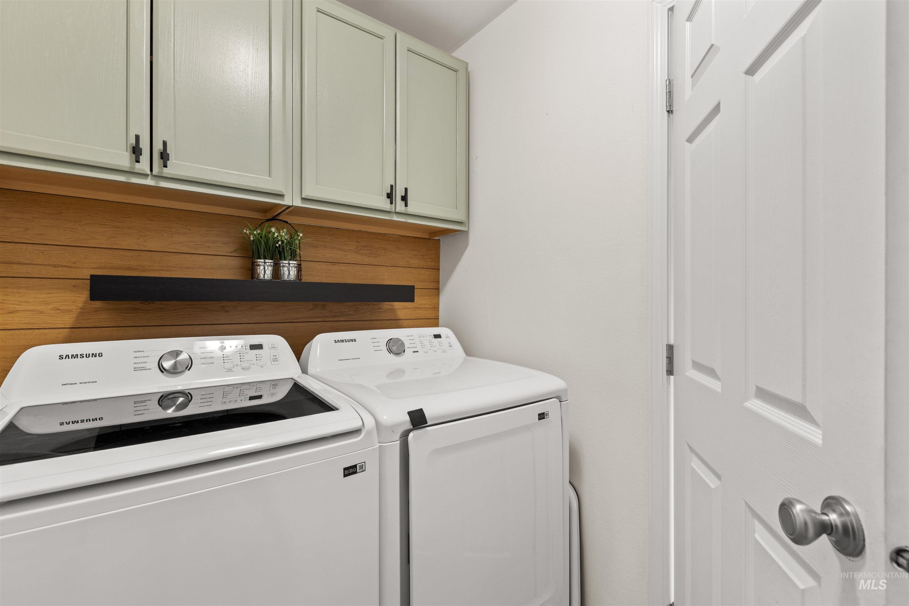 Dedicated laundry area featuring light green upper cabinetry with dark hardware, a wood-finish shiplap accent wall, and a matte black floating shelf