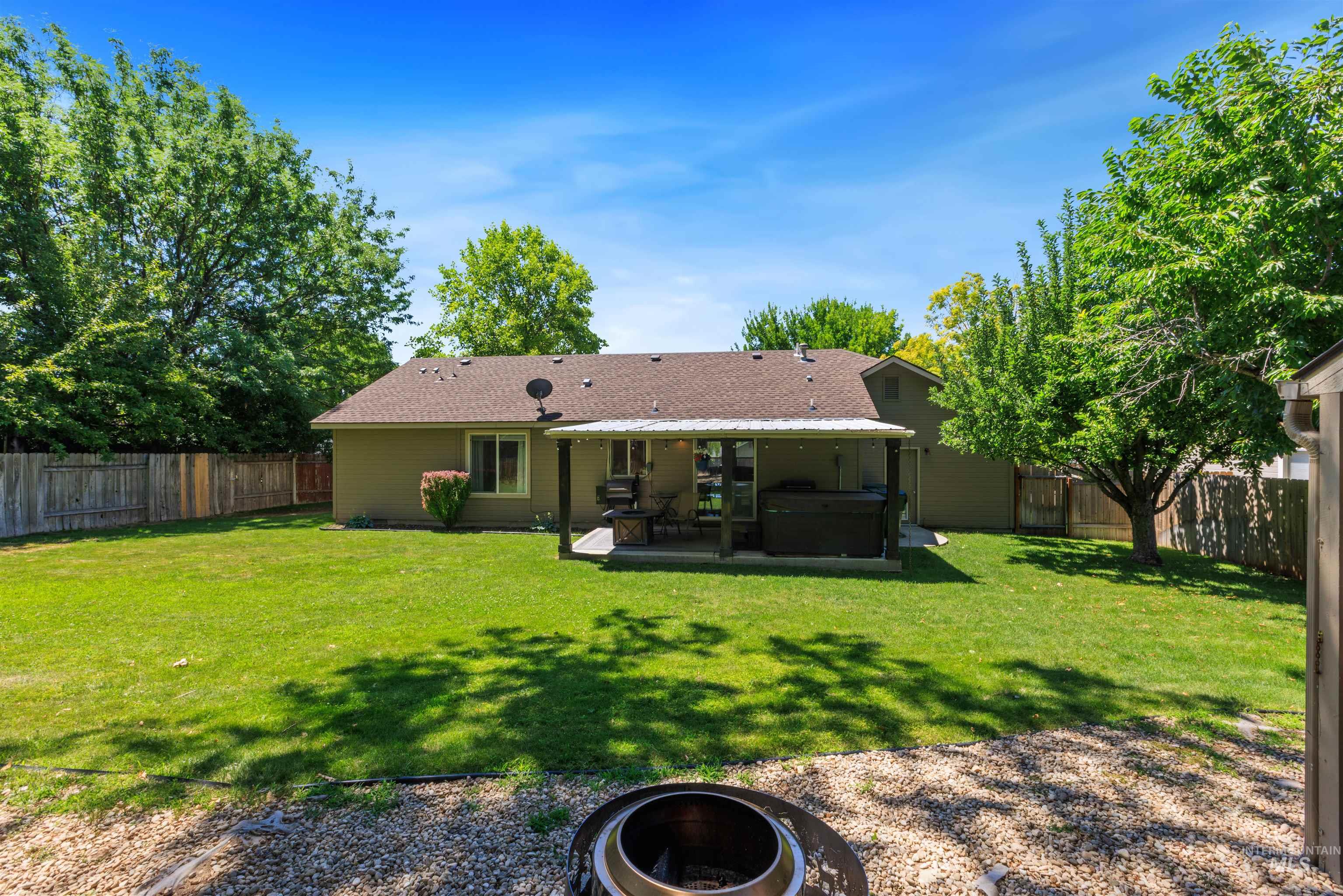 Expansive backyard featuring a manicured lawn, established trees, full privacy fencing, a covered patio, and a fire pit area with gravel ground cover