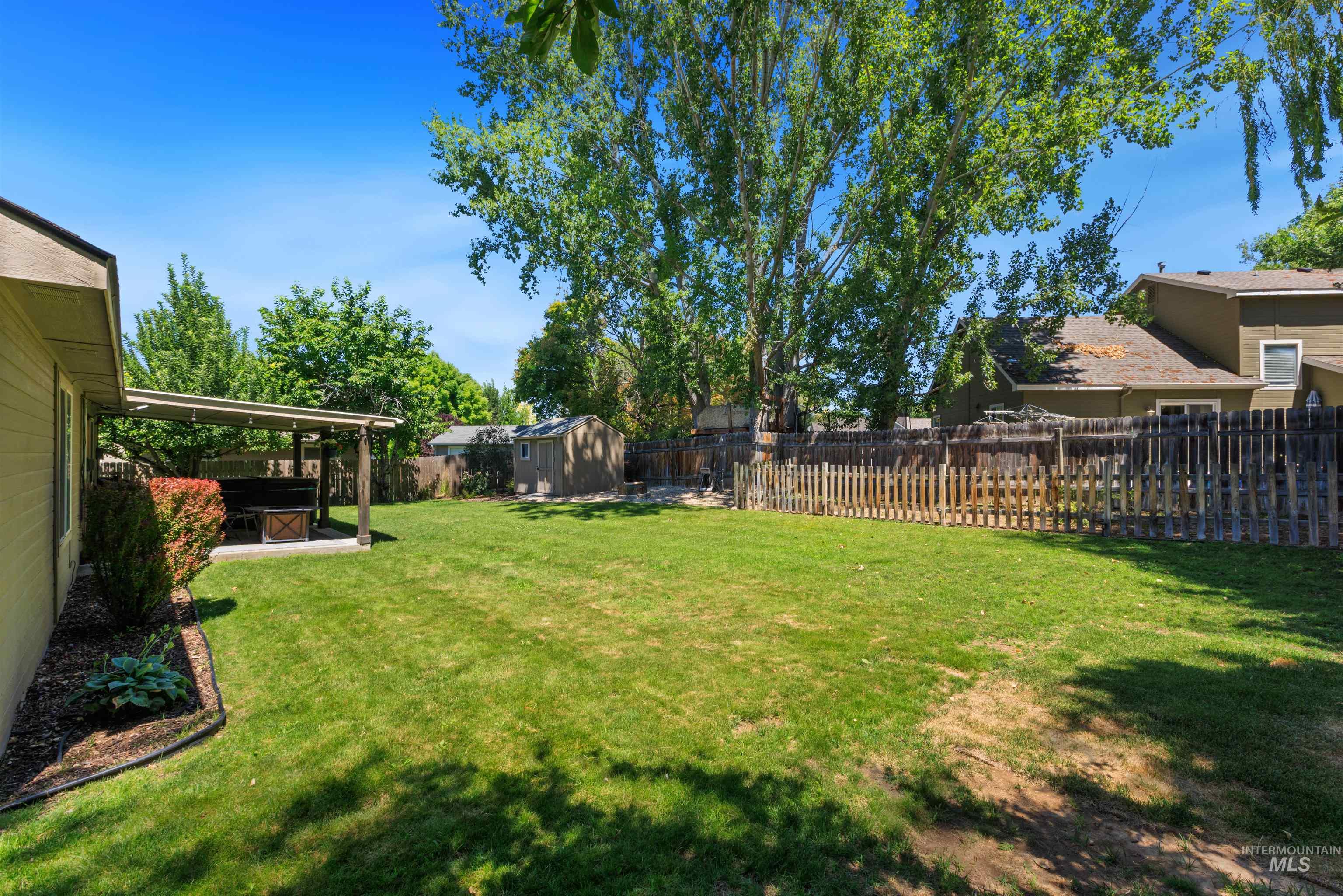 Expansive backyard featuring a covered patio, a detached storage shed, lush green lawn, mature perimeter trees, and a full privacy fence