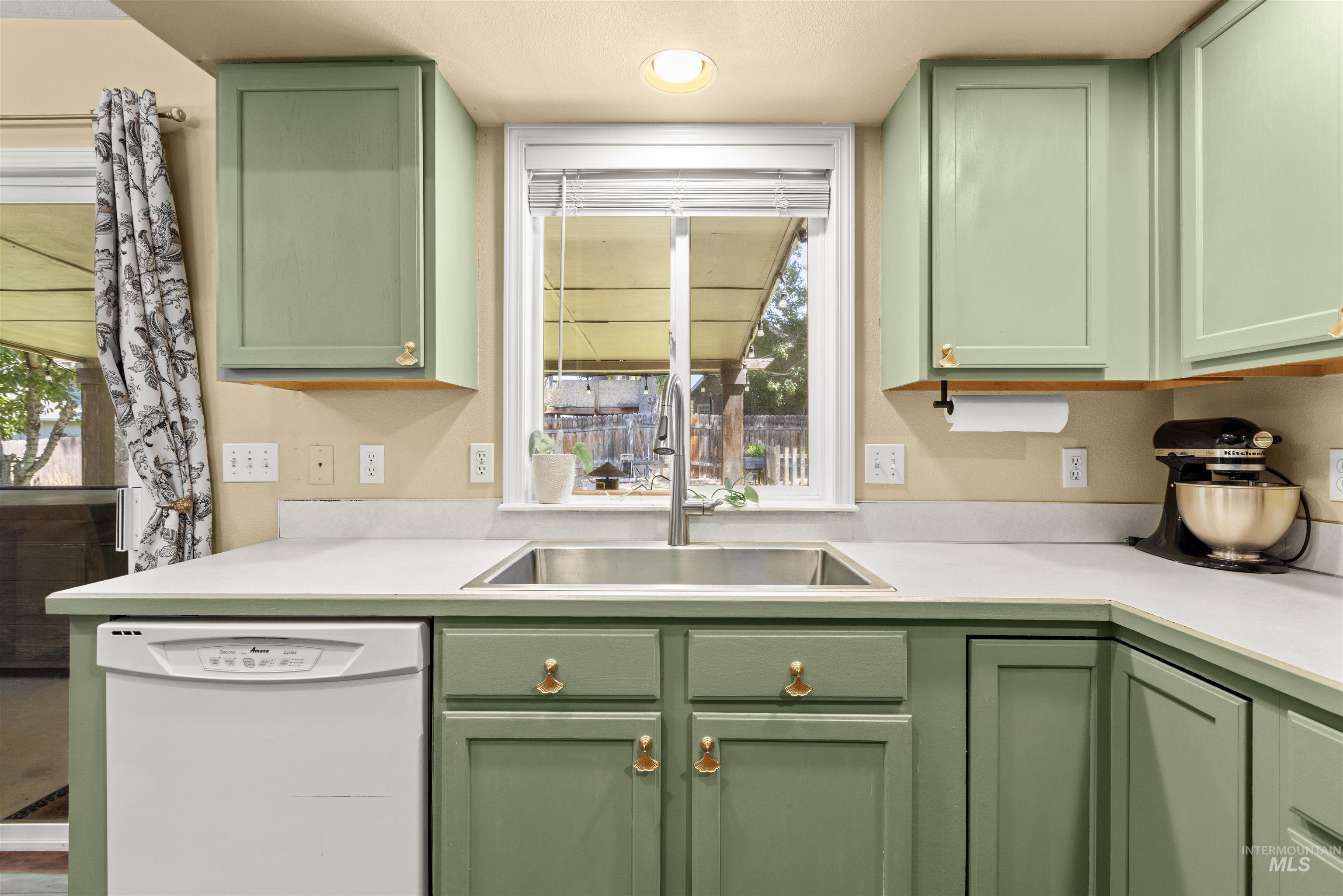 Kitchen featuring sage green cabinetry with gold-toned hardware, light countertops, a stainless steel sink with a gooseneck faucet, a white dishwasher, and a window with blinds overlooking the exterior