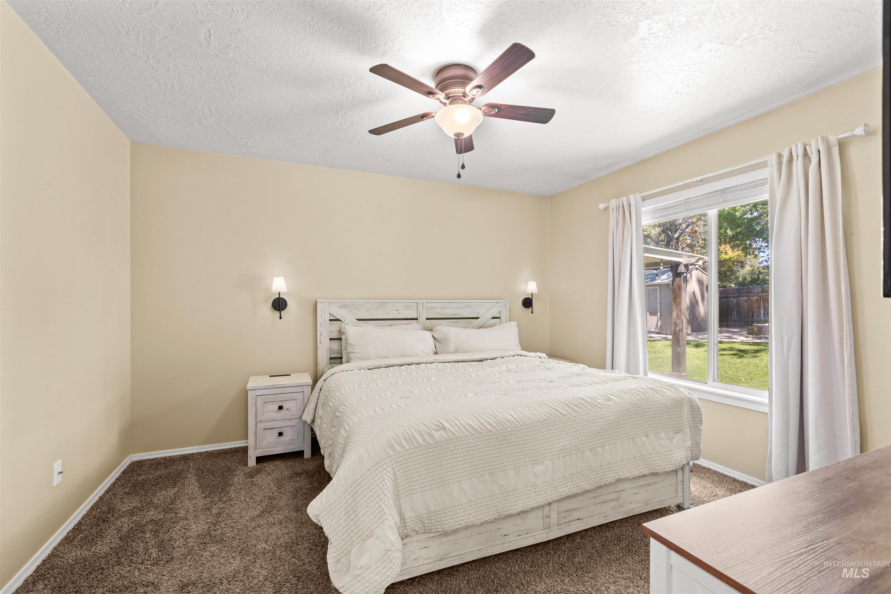 Bright bedroom featuring a large window, ceiling fan with integrated lighting, wall-mounted sconces, and neutral-toned carpet flooring