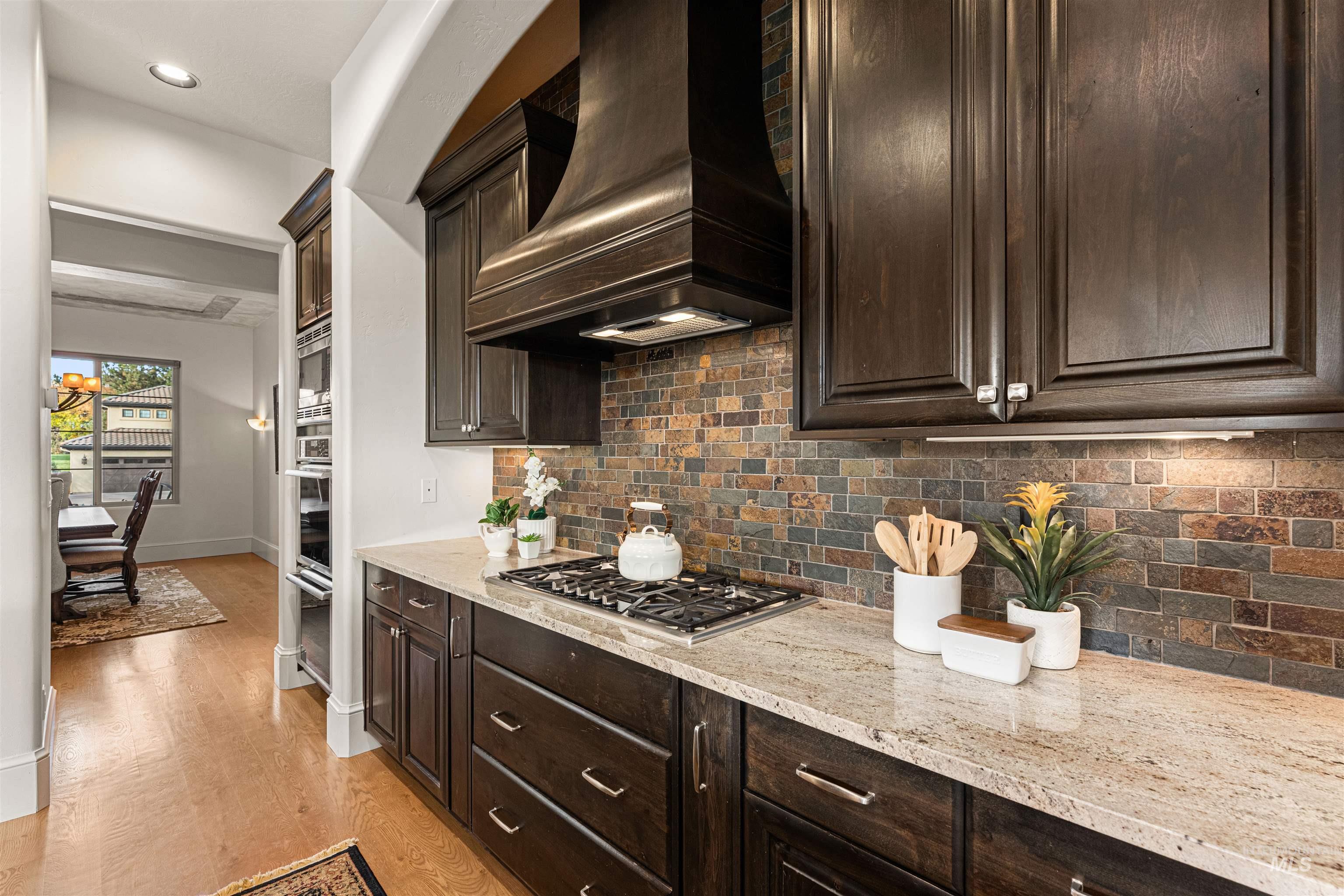 Kitchen with dark brown cabinets, decorative backsplash, light stone countertops, custom exhaust hood, and stainless steel appliances