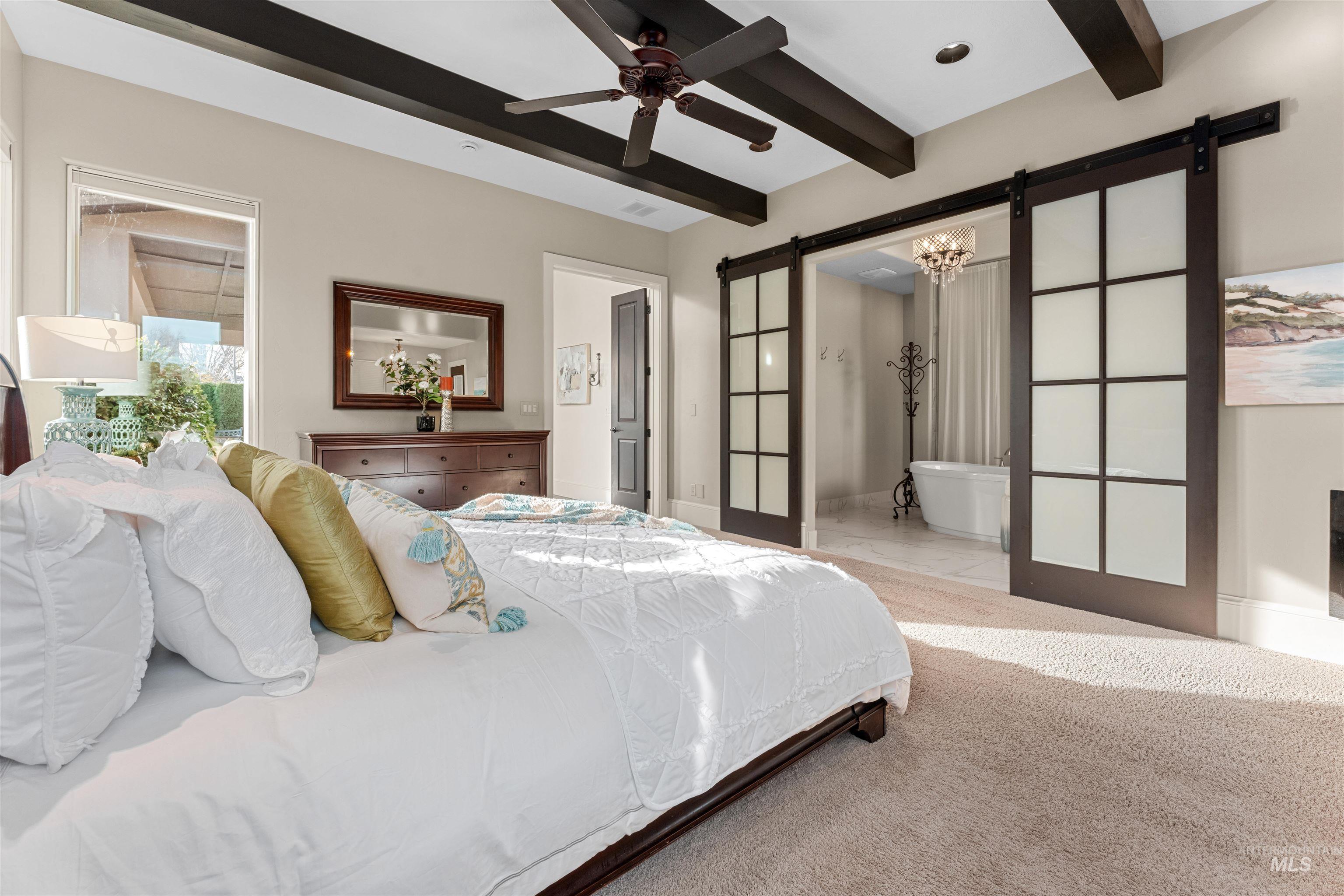 Carpeted bedroom featuring a barn door, beamed ceiling, ceiling fan, ensuite bathroom, and a chandelier