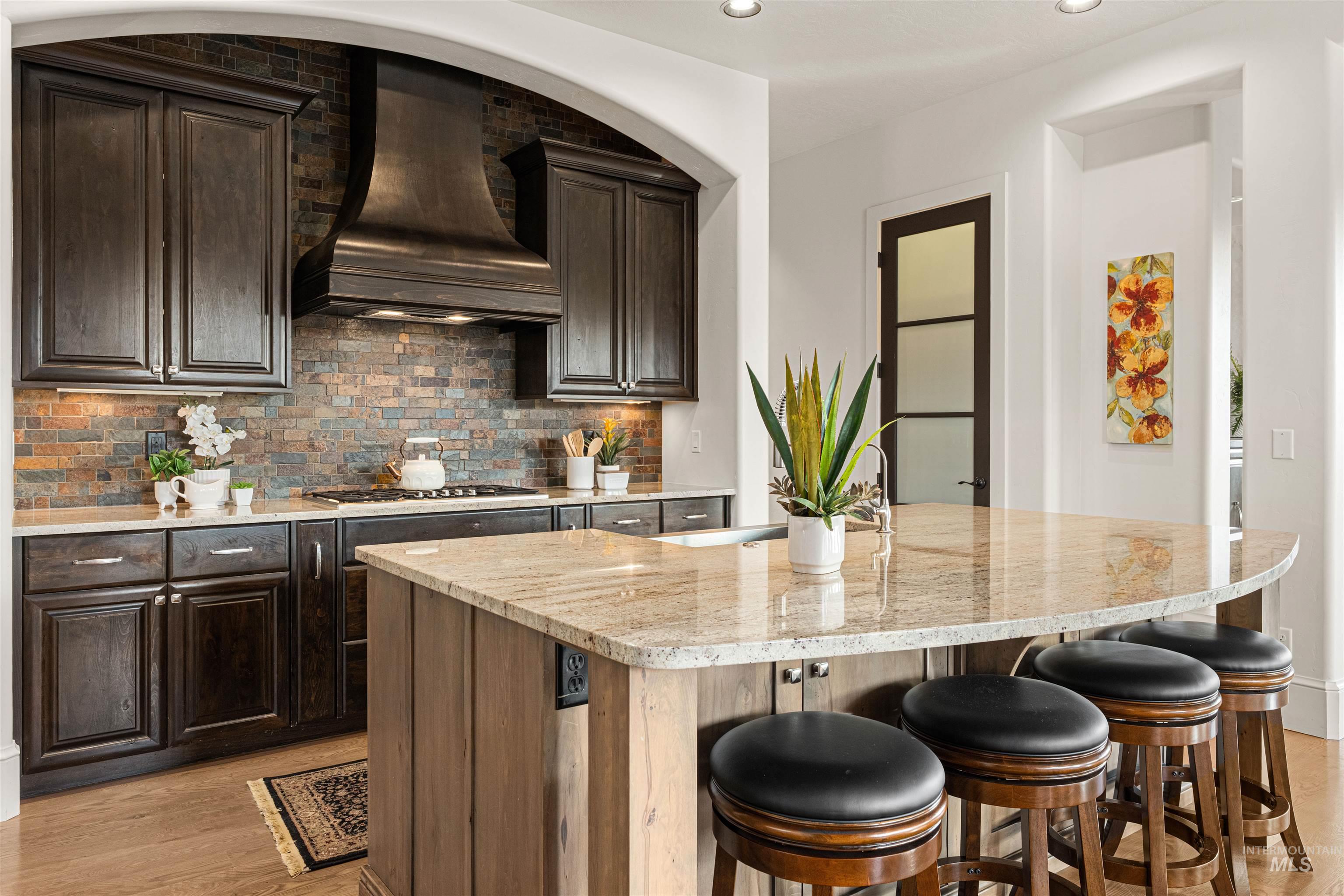 Kitchen with a kitchen bar, backsplash, light stone countertops, dark brown cabinets, and custom range hood