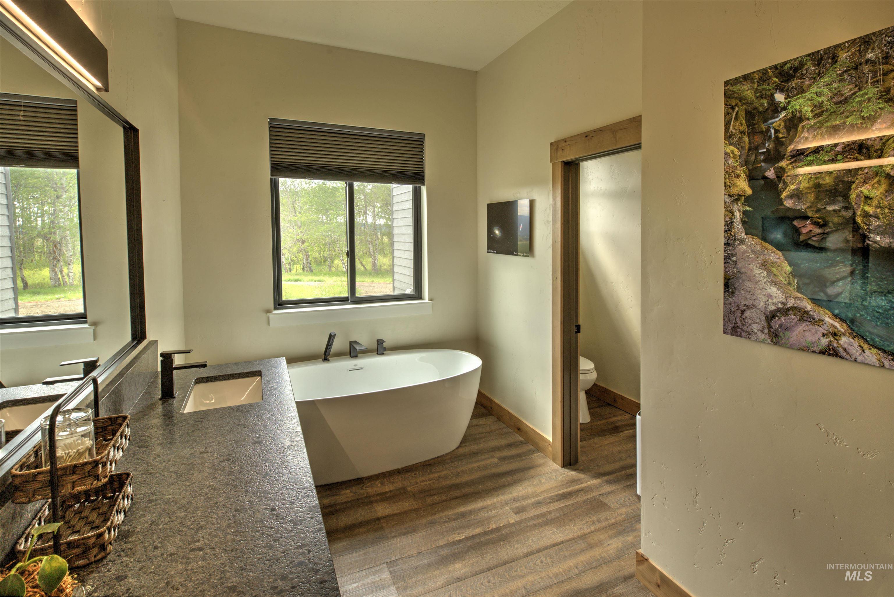 Freestanding soaking tub with dark fixtures, dual windows, wood-finish flooring, and a stone-finish countertop with an integrated sink