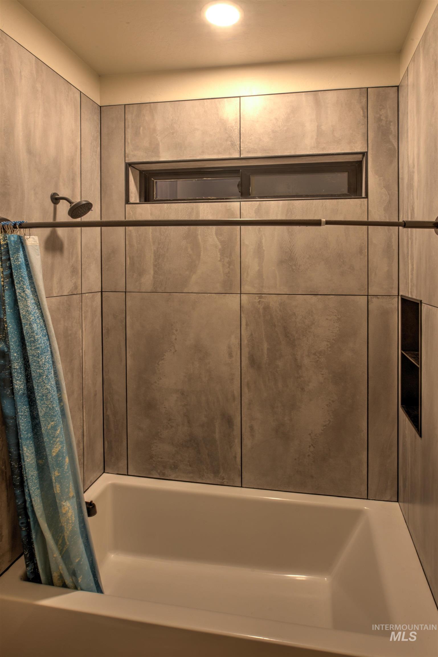 Bathtub with floor-to-ceiling tile surround, featuring a built-in shower niche, a high transom window, and a matte black shower head