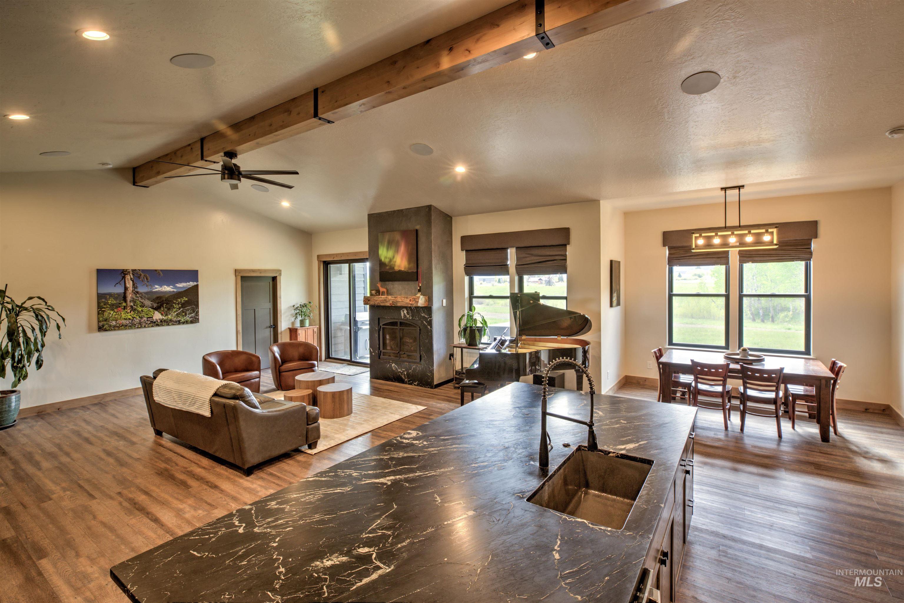 Open-concept interior featuring wood-finish flooring, exposed ceiling beams, recessed lighting, a stone-clad fireplace, and a kitchen island with a dark countertop and integrated sink