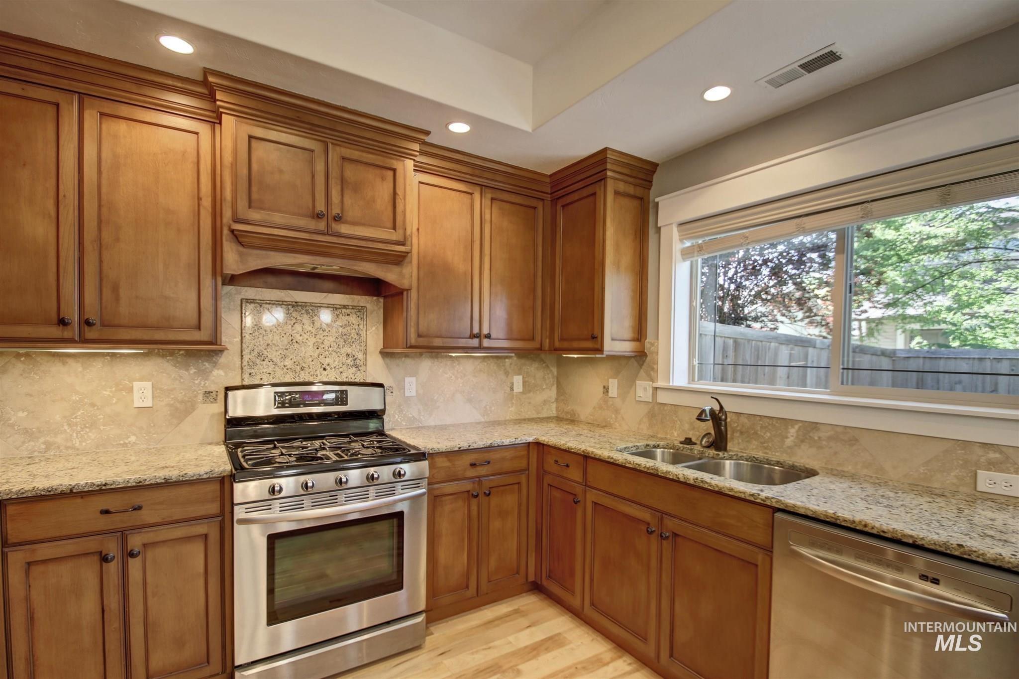 Kitchen with stainless steel appliances, brown cabinetry, light stone counters, recessed lighting, and light wood-style floors