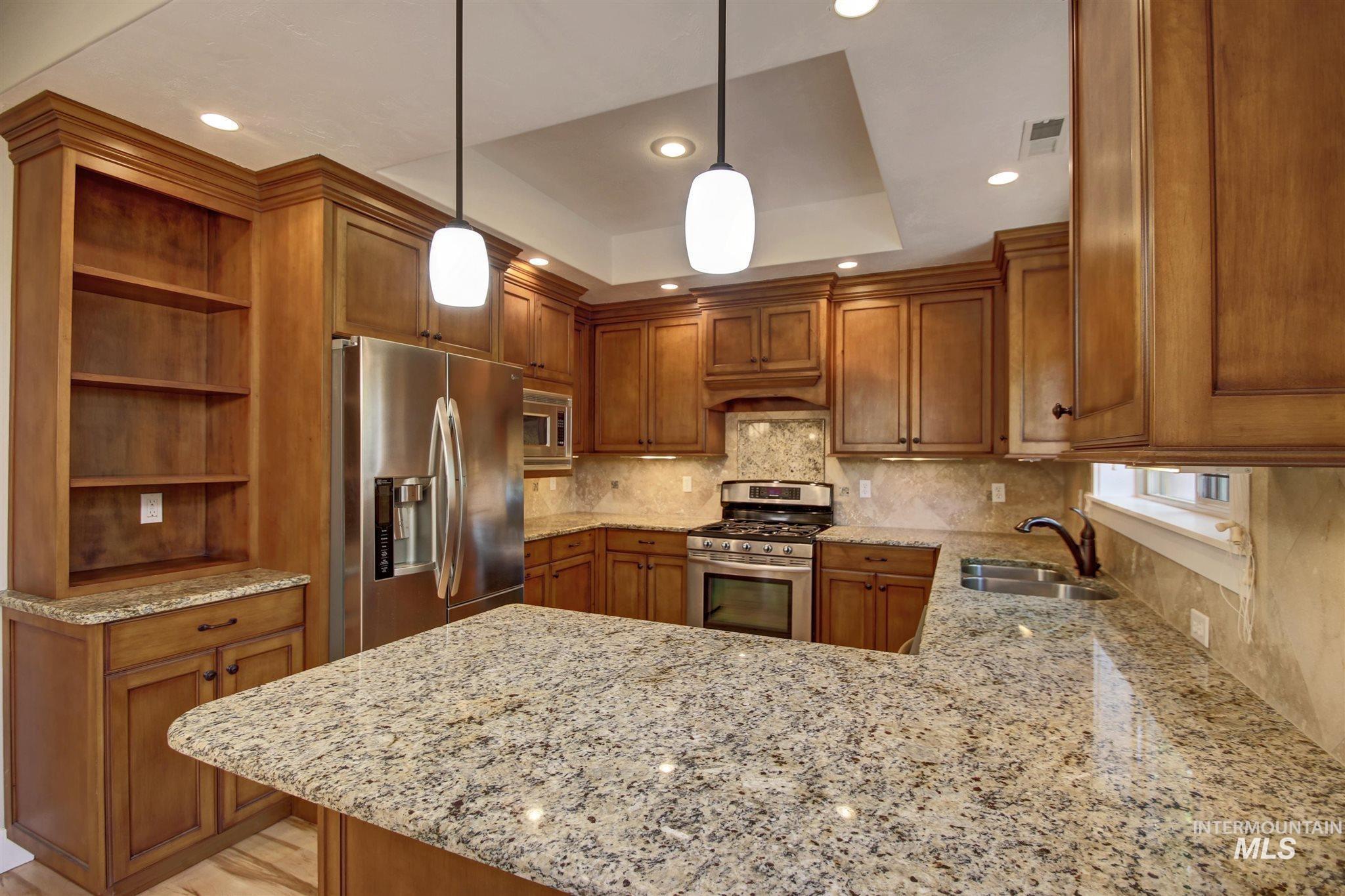 Kitchen featuring brown cabinetry, light stone countertops, appliances with stainless steel finishes, a peninsula, and a tray ceiling