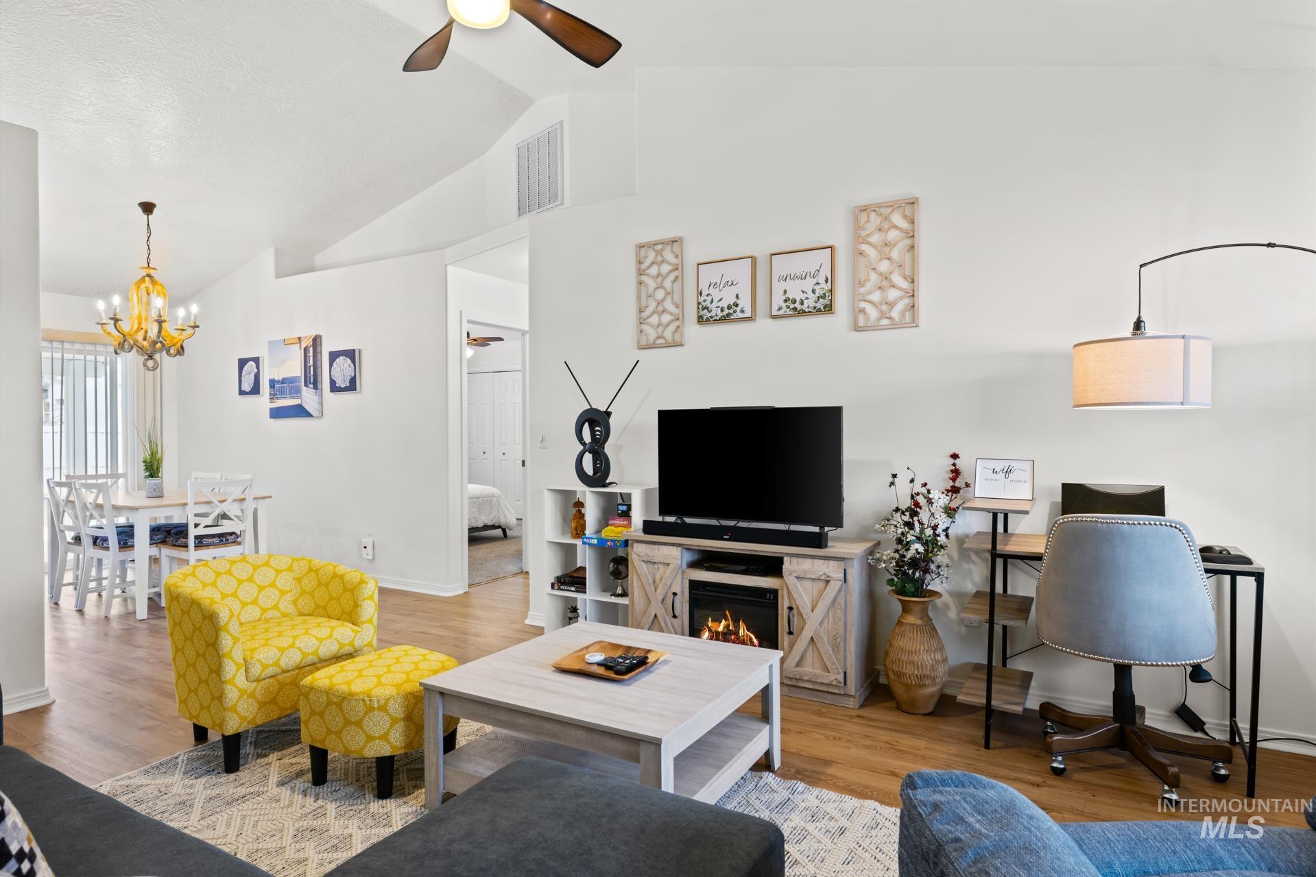 Living area featuring wood finished floors, ceiling fan, vaulted ceiling, a chandelier, and a warm lit fireplace