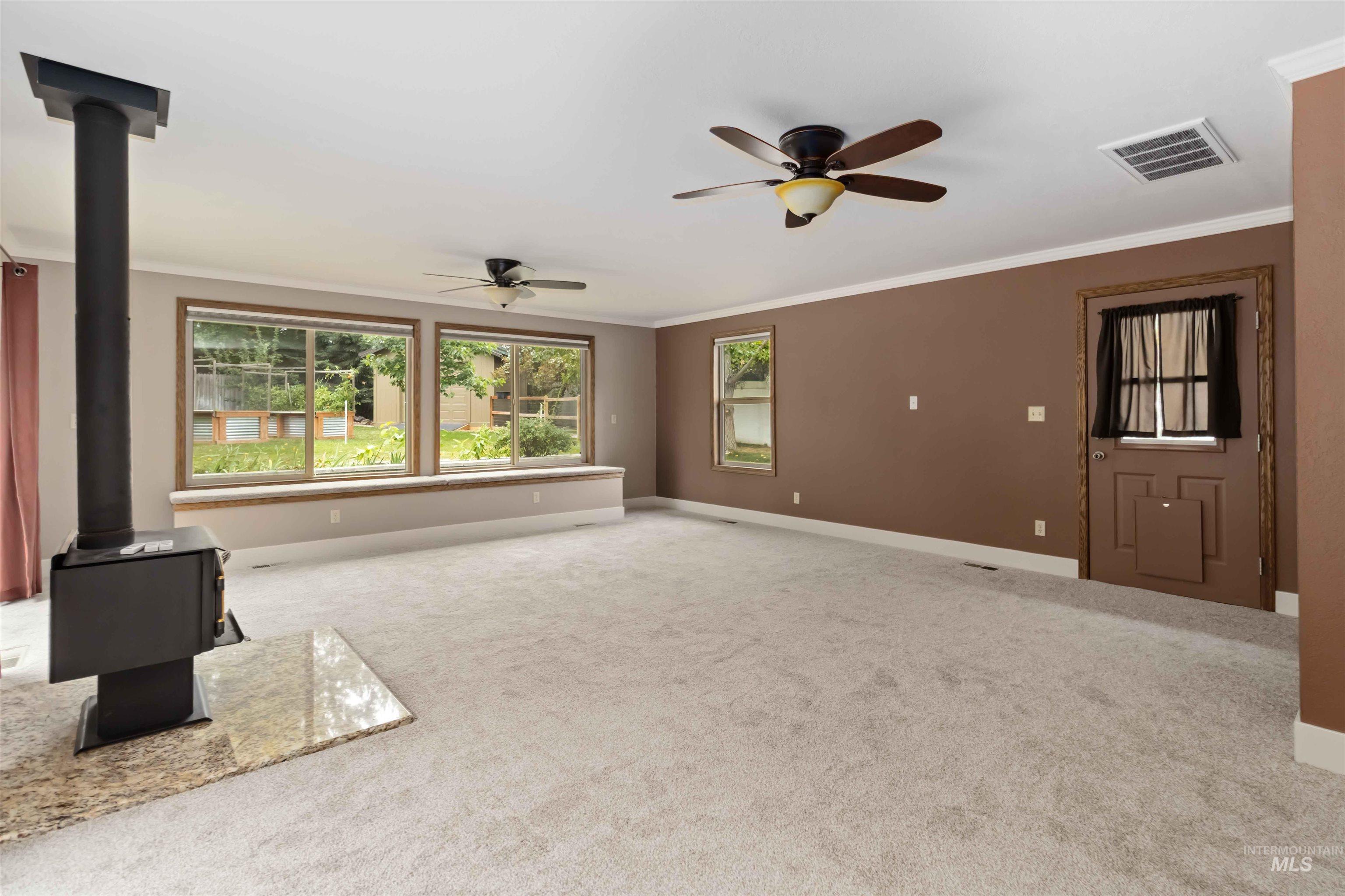 Unfurnished living room featuring a wood stove, crown molding, carpet flooring, and ceiling fan