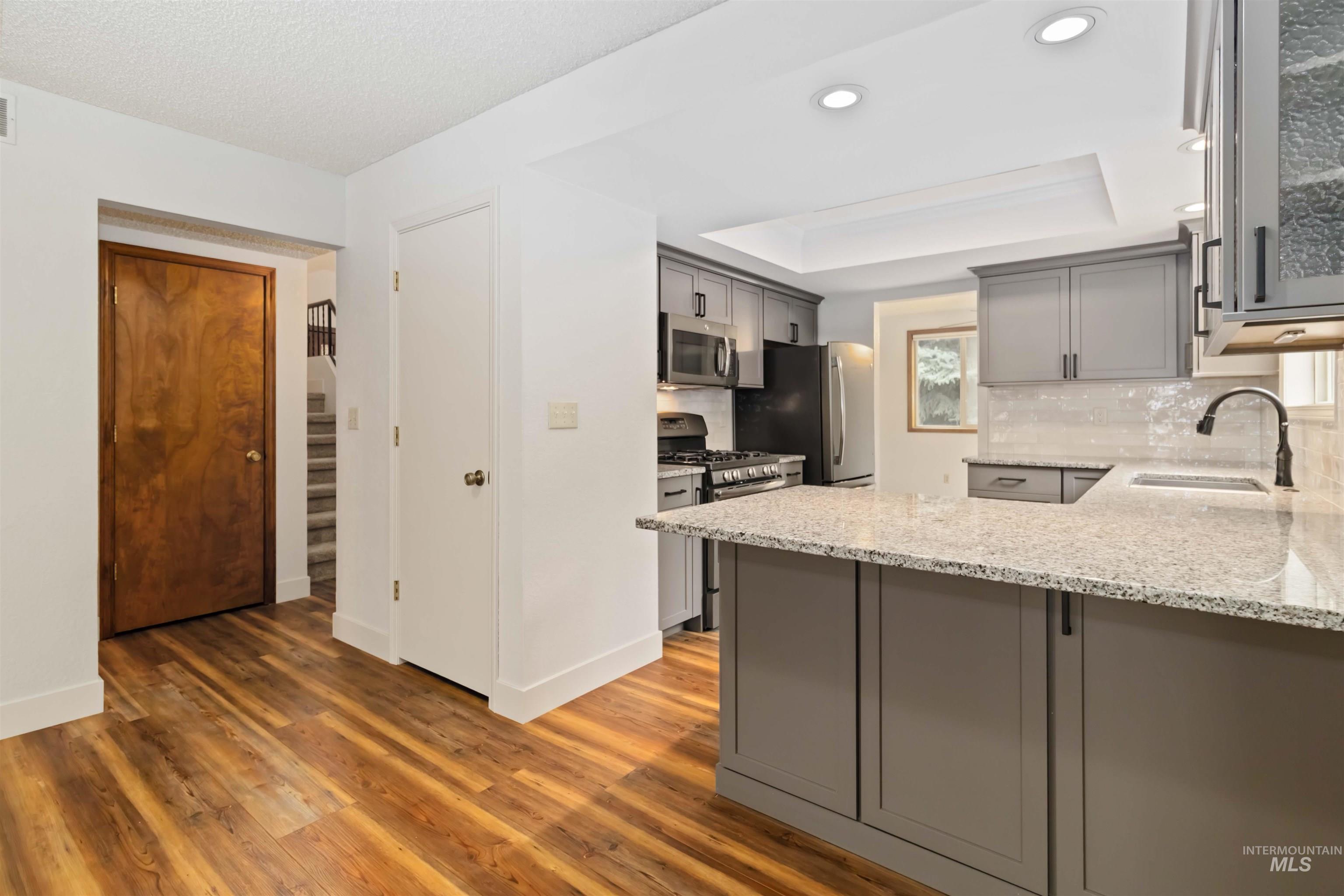 Kitchen with gray cabinets, light stone countertops, a raised ceiling, appliances with stainless steel finishes, and tasteful backsplash