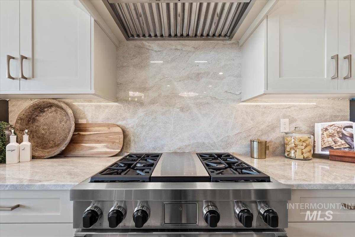 Kitchen with custom range hood, tasteful backsplash, light stone countertops, stainless steel gas stovetop, and white cabinetry