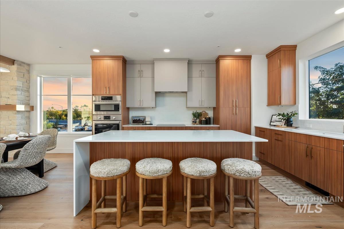 Modern kitchen featuring a waterfall island with an integrated breakfast bar, stainless steel built-in appliances, a range hood, and a subway tile backsplash
