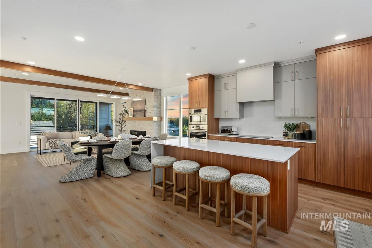 Open-concept living area featuring light wood-finish flooring, a contemporary kitchen with a large island and integrated appliances, exposed wood beams, and a stone-clad fireplace