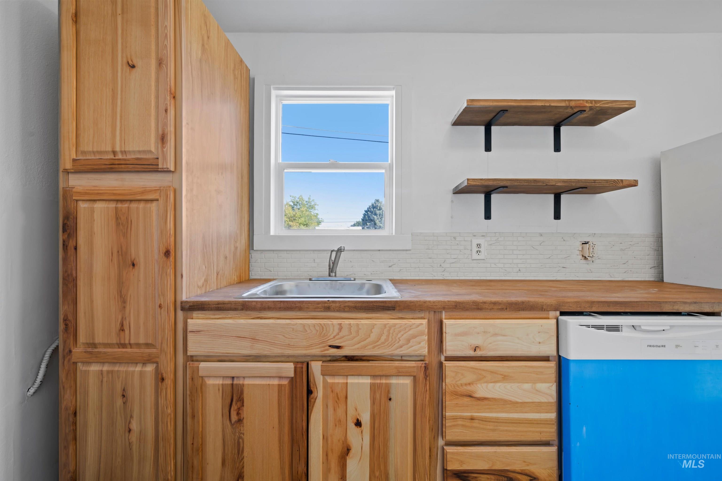 Kitchen featuring white dishwasher, backsplash, light brown cabinetry, open shelves, and wood counters