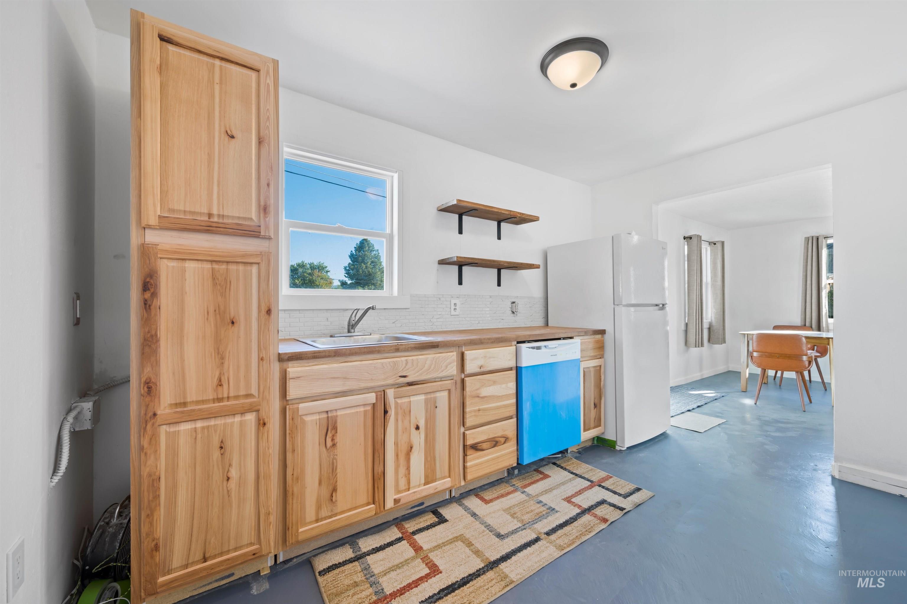 Kitchen with light brown cabinetry, backsplash, open shelves, finished concrete flooring, and dishwasher