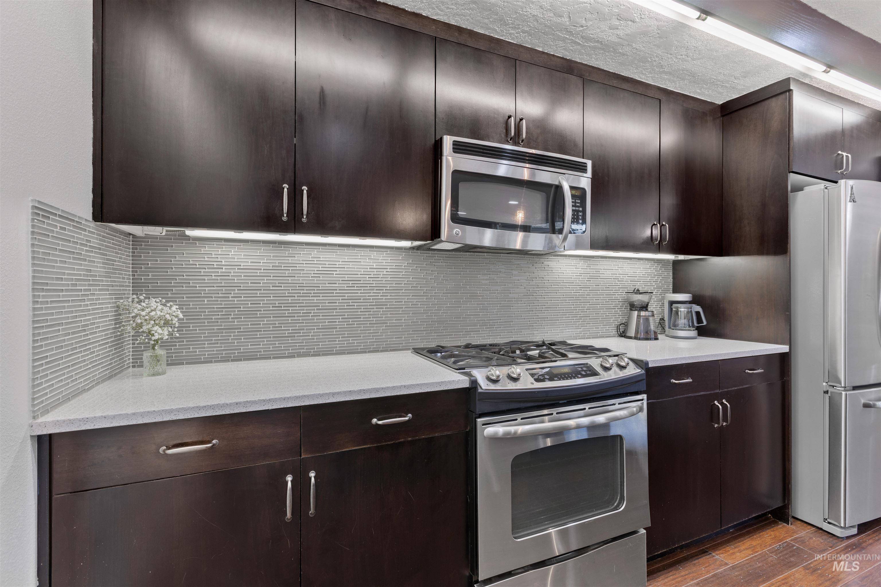 Kitchen with appliances with stainless steel finishes, tasteful backsplash, dark brown cabinets, dark wood-type flooring, and light stone countertops